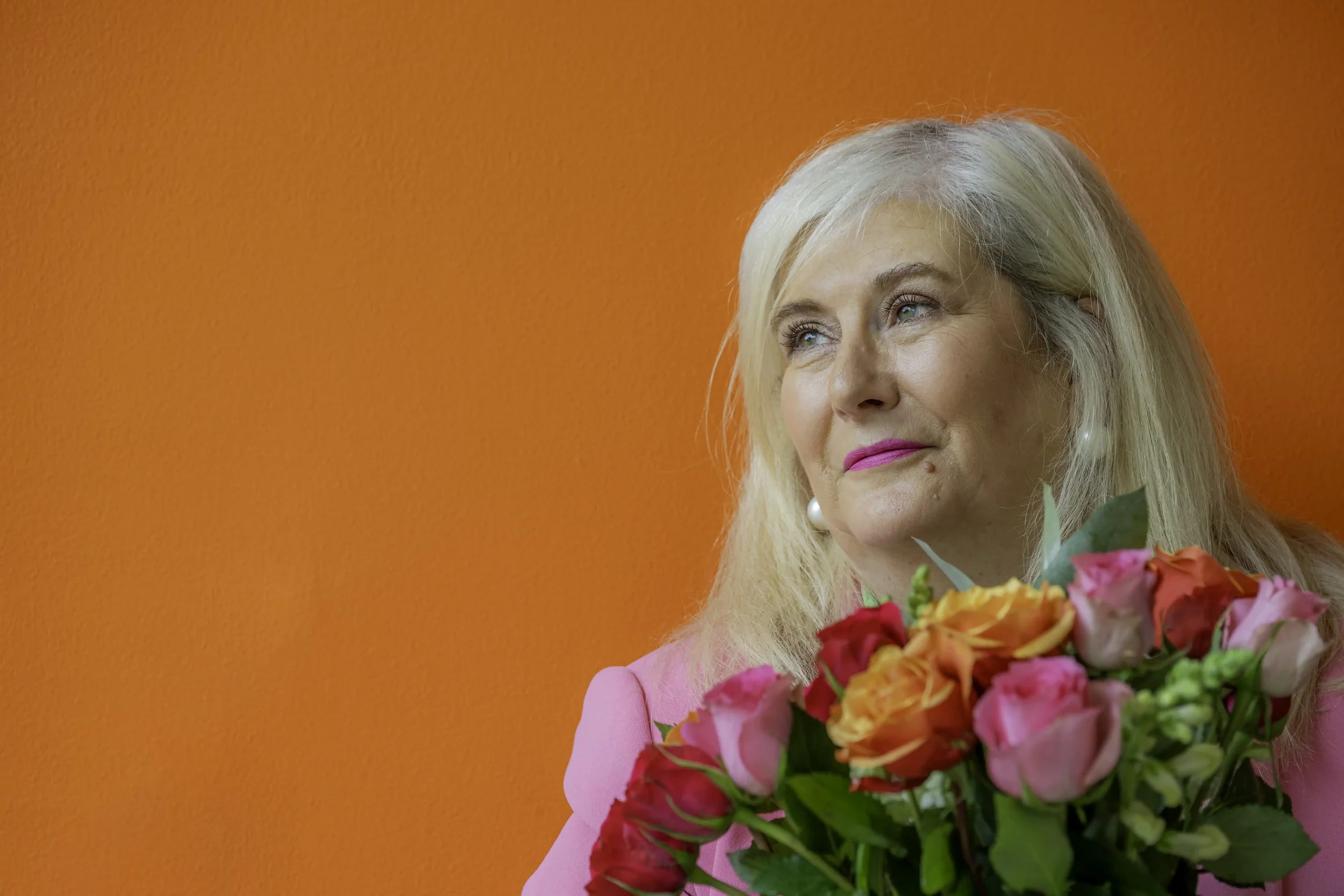 Headshot photograph of an older woman with long blonde hair wearing bright pink lipstick and a pink jacket, holding a bouquet of flowers, standing against a bright oragne wall, not looking directly at the camera