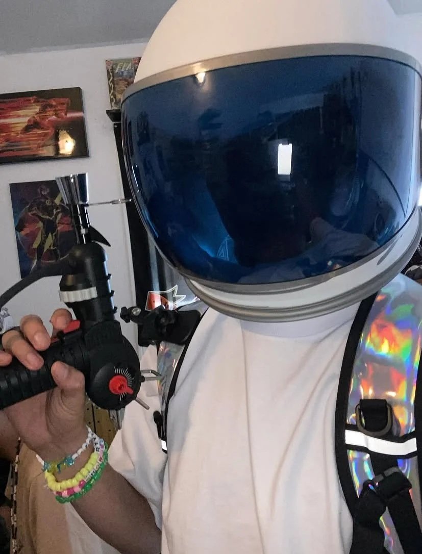 Person wearing an astronaut helmet and holding a paintball gun in a room with posters on the wall.