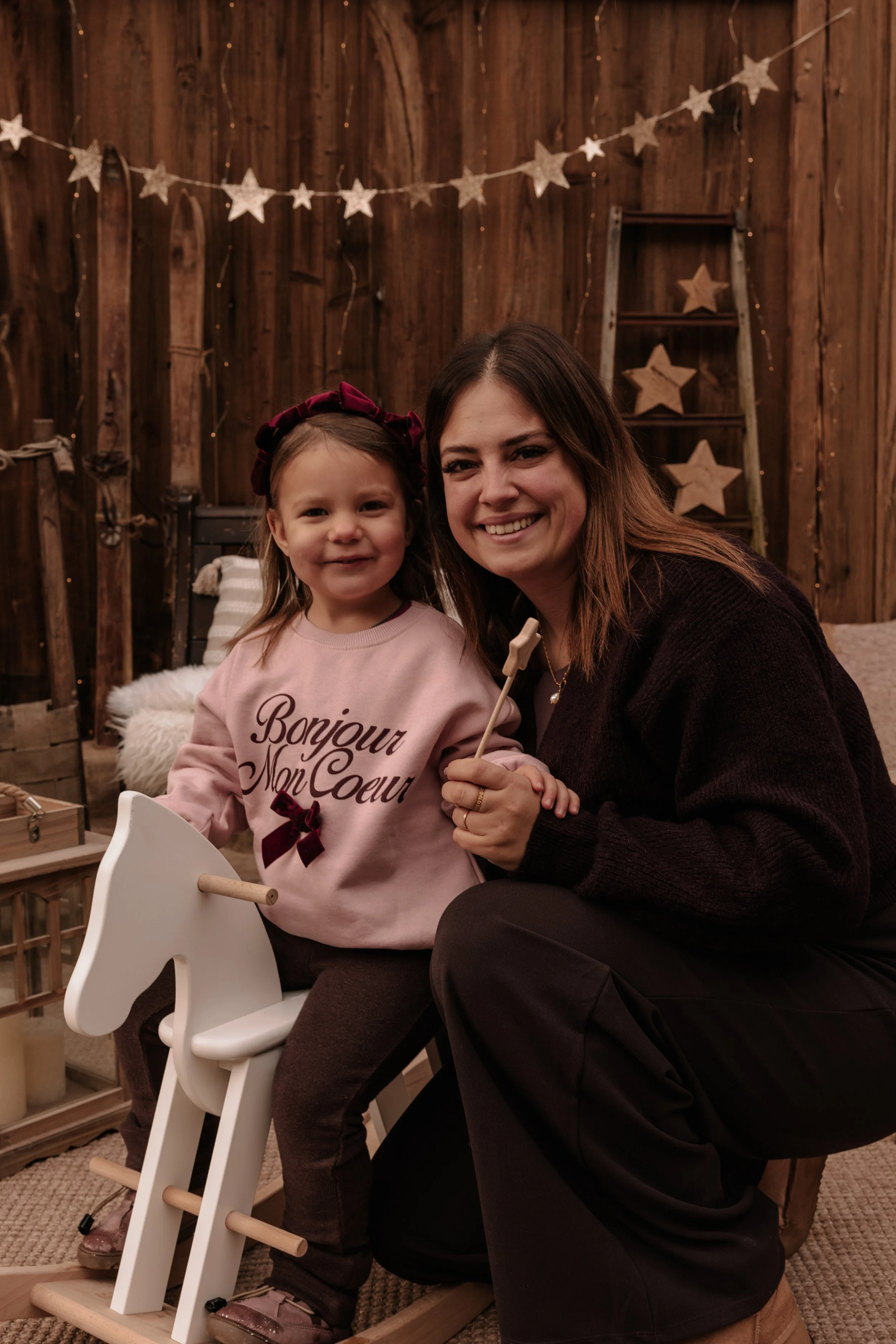 Eine junge Frau und ein kleines Mädchen sitzen in einem rustikalen Raum mit Holzvertäfelung, Sterne und Lichter in der Dekoration. Das Mädchen sitzt auf einem weißen Holzpferd und trägt ein pinkfarbenes Sweatshirt mit der Aufschrift ‚Bonjour Mon Coeu