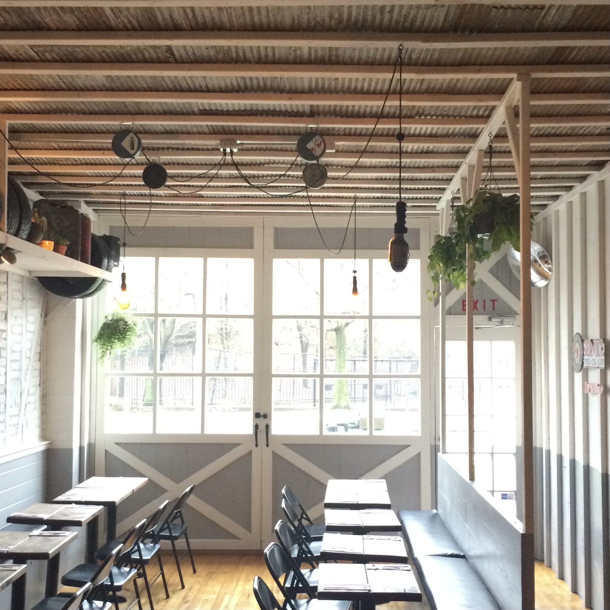 Interior of a rustic restaurant with wooden tables, black chairs, and a corrugated metal ceiling. Large windows and hanging plants add a touch of greenery. The exit sign is visible above the door.