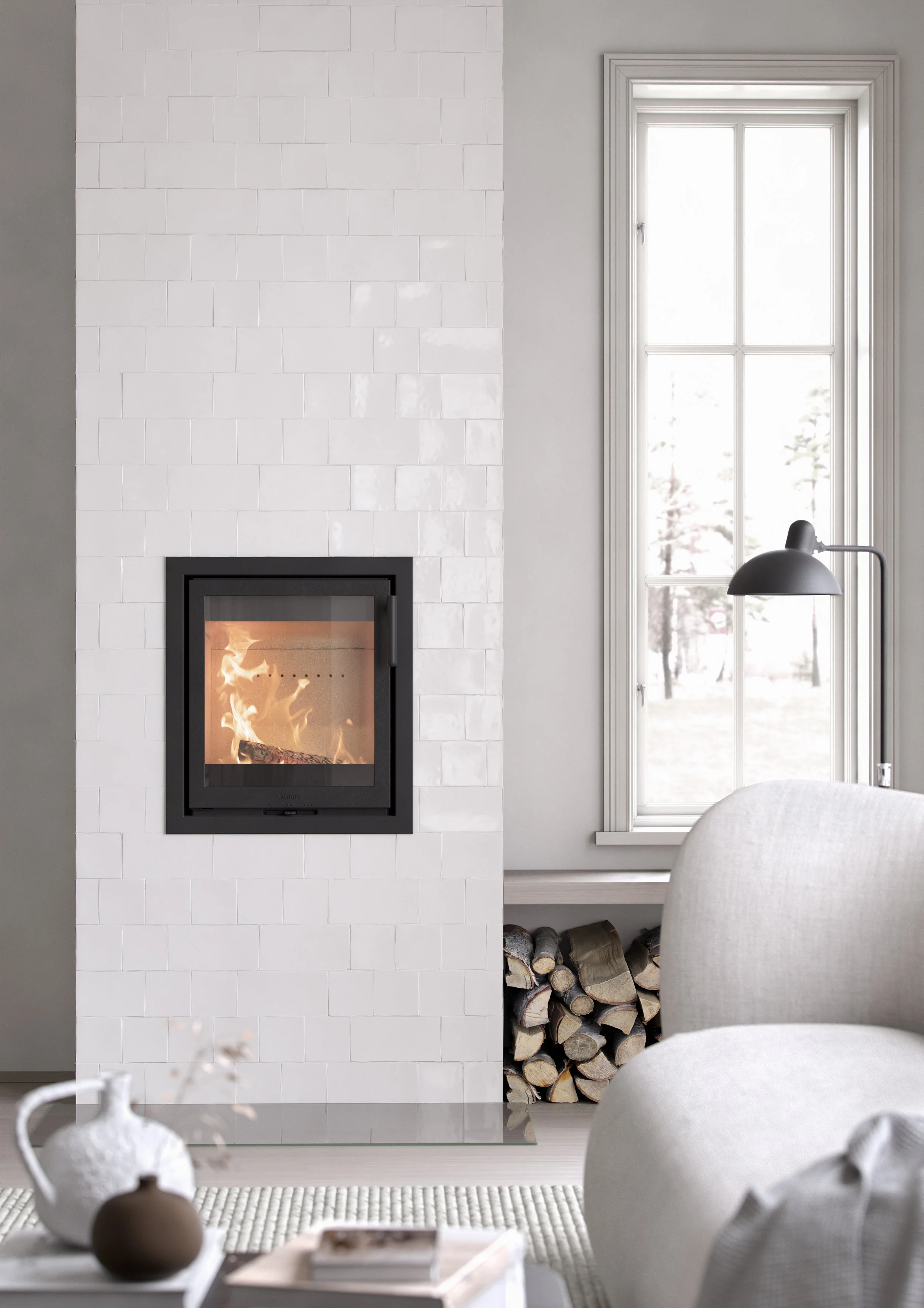 Living room with a fireplace on the left, a large window to the right, and a white armchair in the foreground. There's a stack of firewood under the window and a black floor lamp beside it. The room has white walls and a beige rug.
