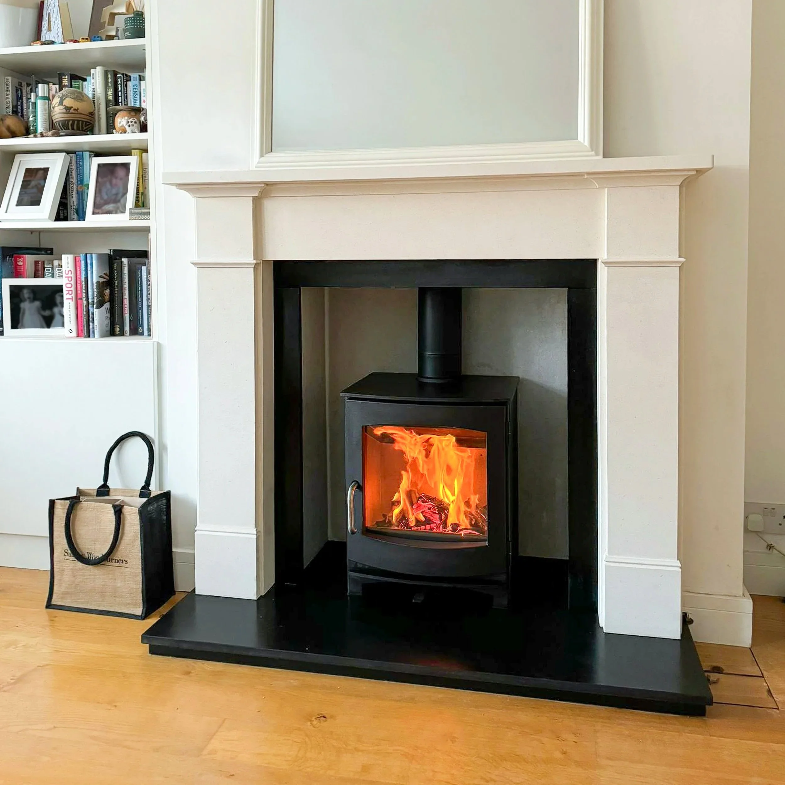 A modern living room with a white fireplace mantel and a black electric fireplace insert displaying flames. To the left, there is a white bookshelf filled with books, framed photos, and decorative items. A beige tote bag with black handles is on the wooden floor in front of the bookshelf.