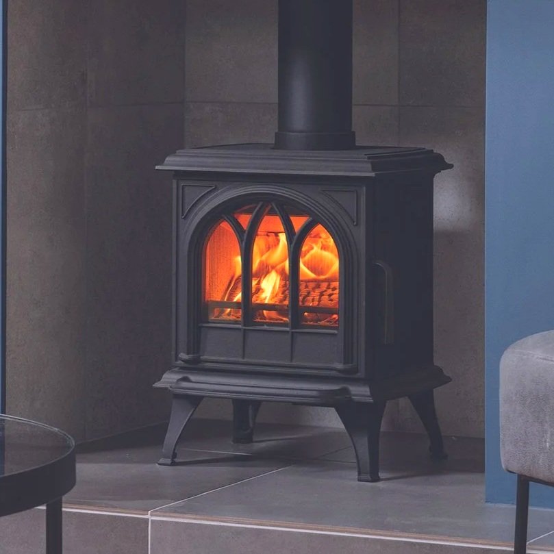 A black wood-burning stove with a glass door showing a fire inside, positioned on a tiled floor against a wall.