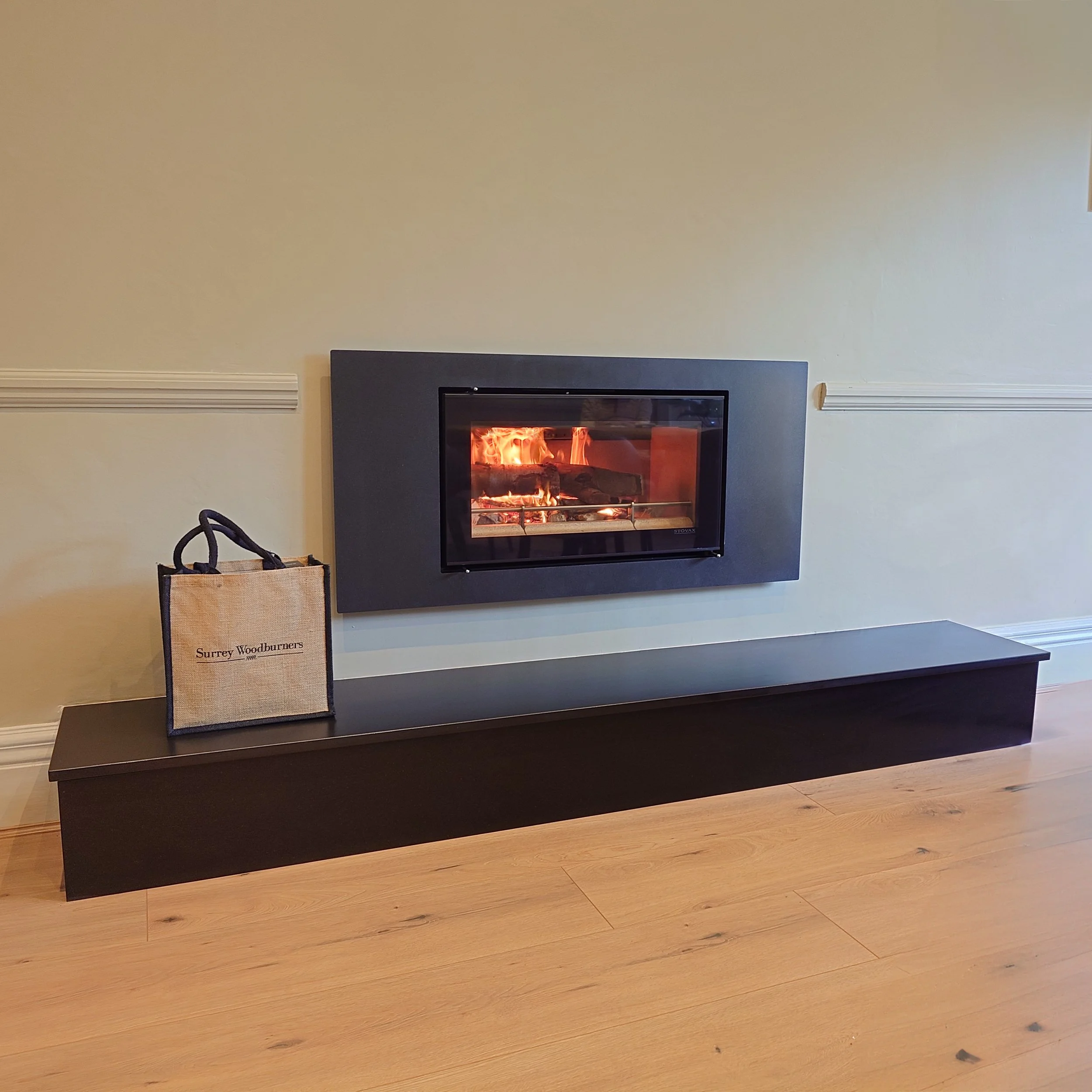 Modern fireplace with flames, black mantel, and a beige bag labeled 'Surrey Woodburners' placed on a black hearth over light wood flooring.