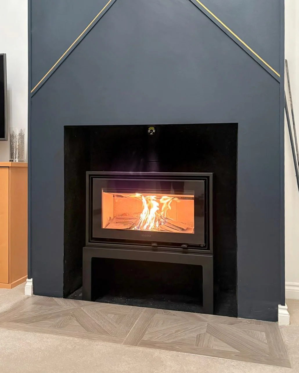 Stovax Studio Air Freestanding Wood Stove installed within an existing modern decorated chimney. The stove sits on a black stove bench. 
