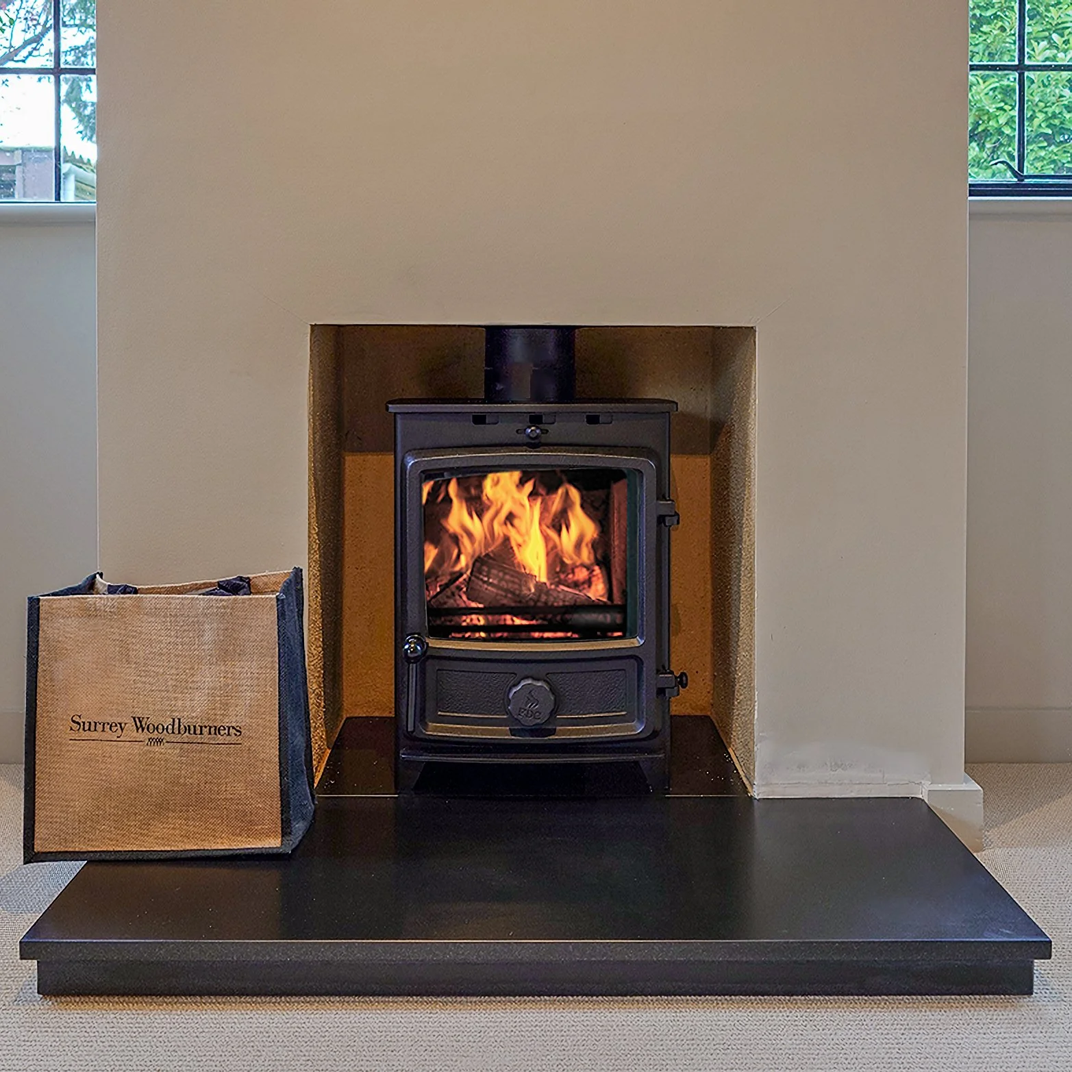 Fire burning inside a black wood stove in a fireplace, with a bag of Surrey Woodburners wood logs nearby.
