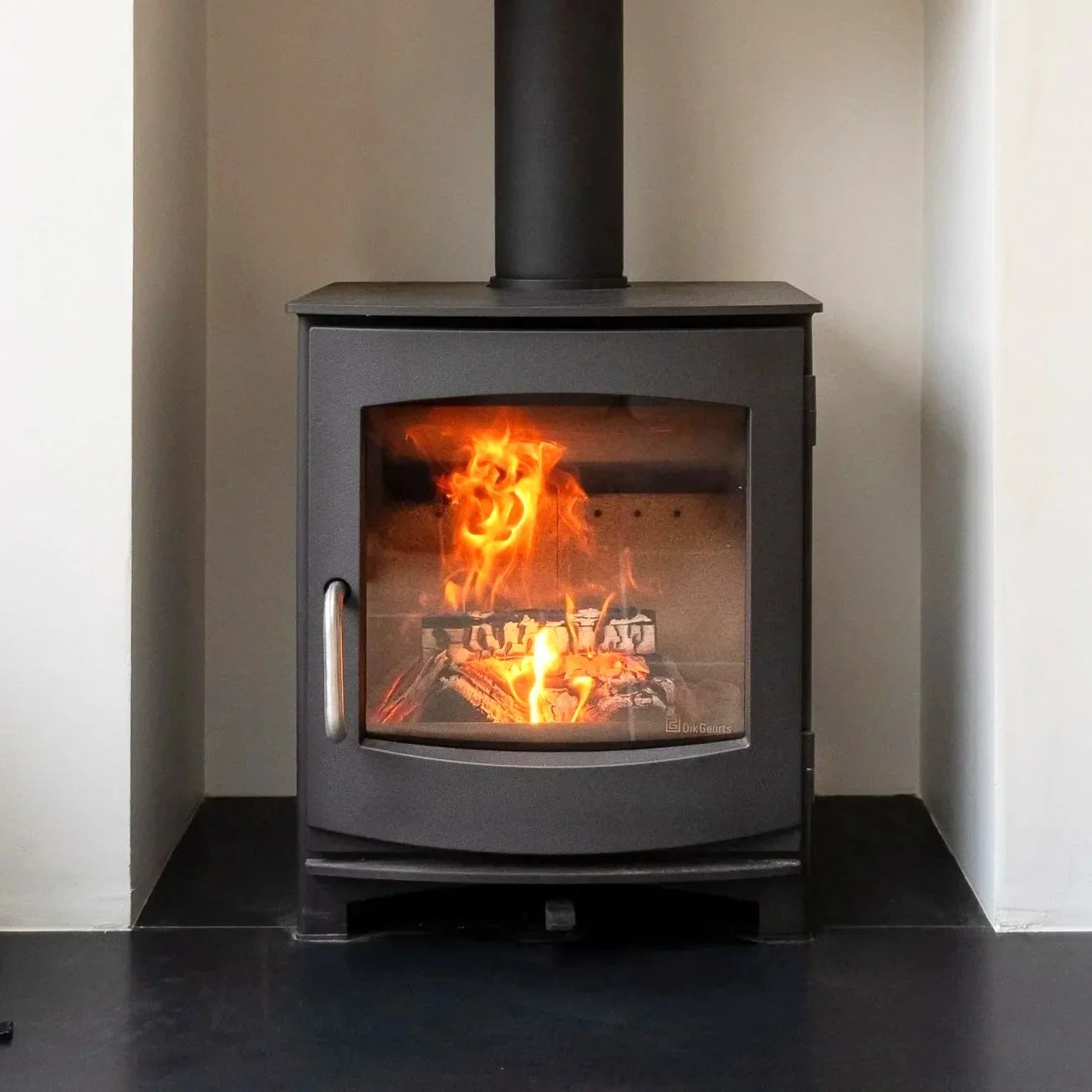 A modern black wood stove with a glass door showing a burning fire inside, situated between two white walls with a dark floor underneath.