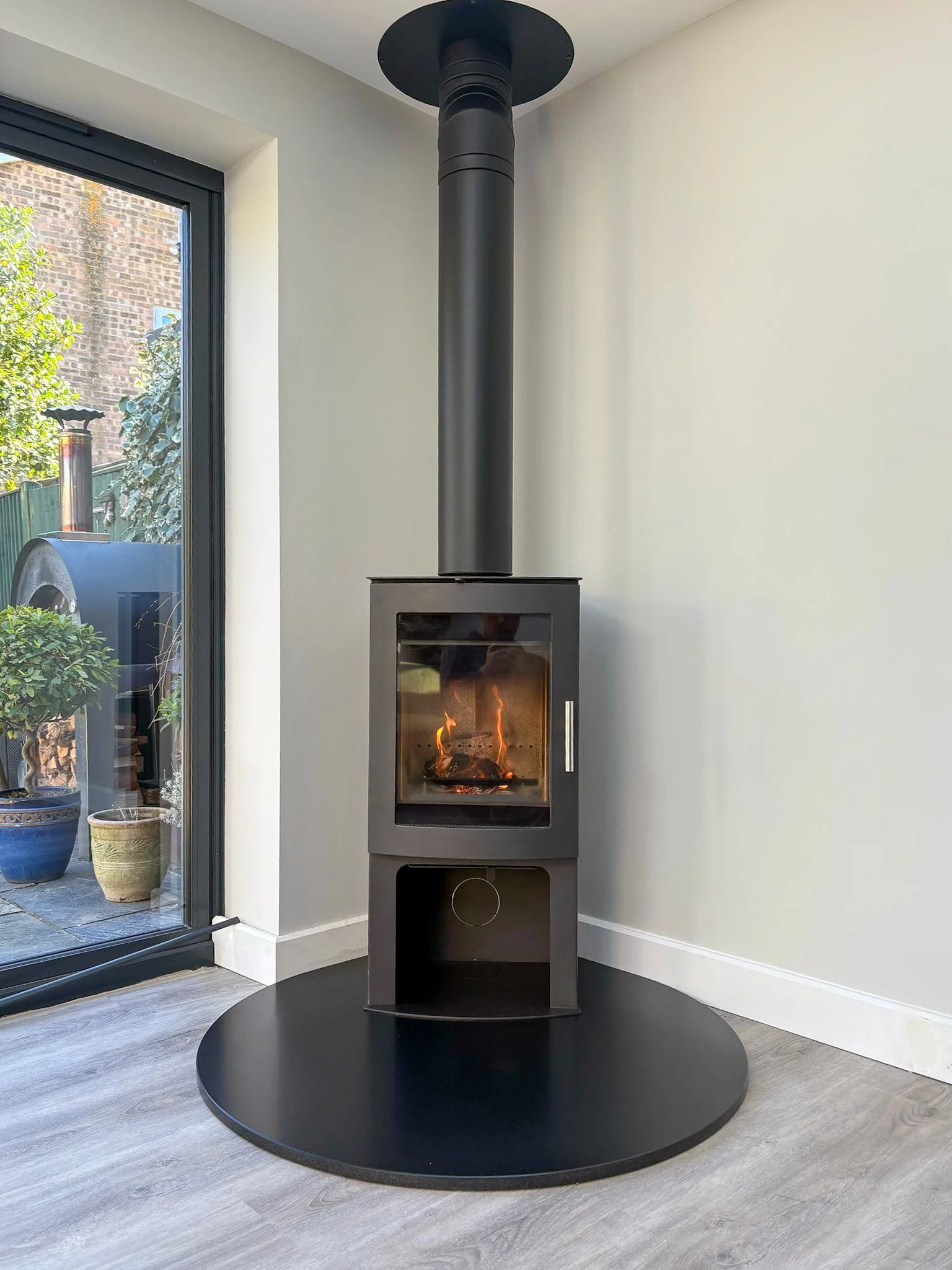 A modern black wood-burning stove with a glass front, showing a fire inside. It sits on a round black platform in a room with light-colored walls and a gray wood floor. A sliding glass door to the left opens to a patio with potted plants and outdoor 