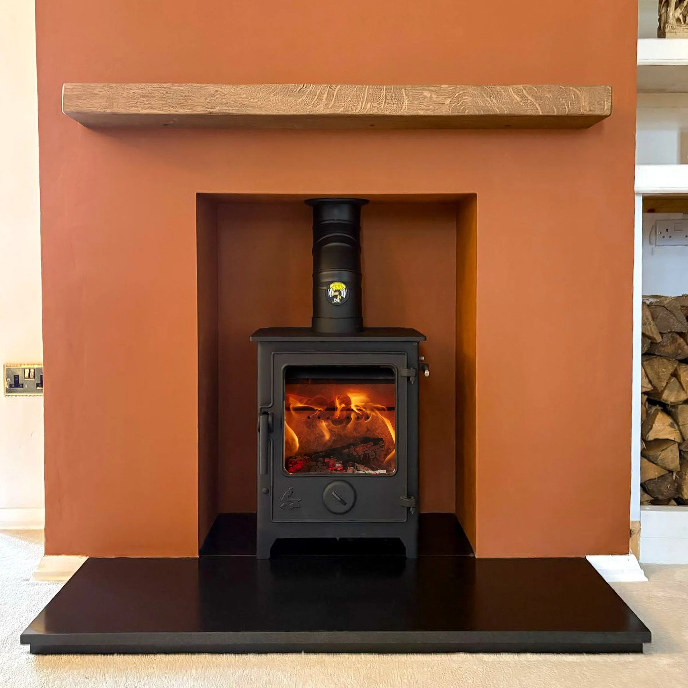 A black wood-burning stove with a glass door showing flames inside, installed in a recessed niche in a burnt orange wall, with a wooden shelf above and a black hearth below.