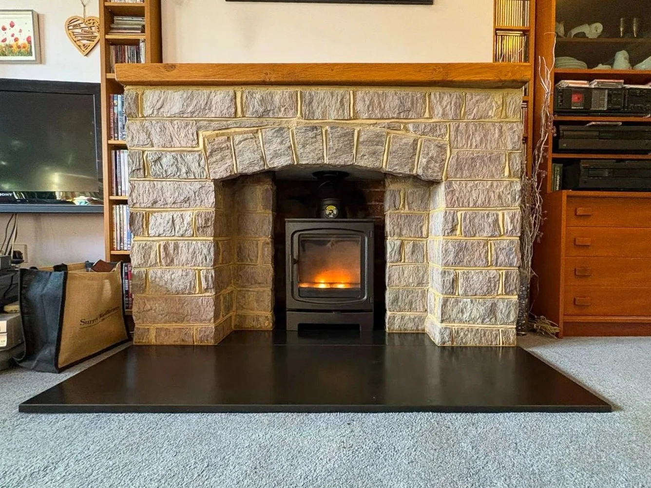 A living room fireplace with a stone surround, a wood mantel, and a black stove with a fire inside. Surrounding the fireplace are bookshelves, a TV, and other electronic equipment.