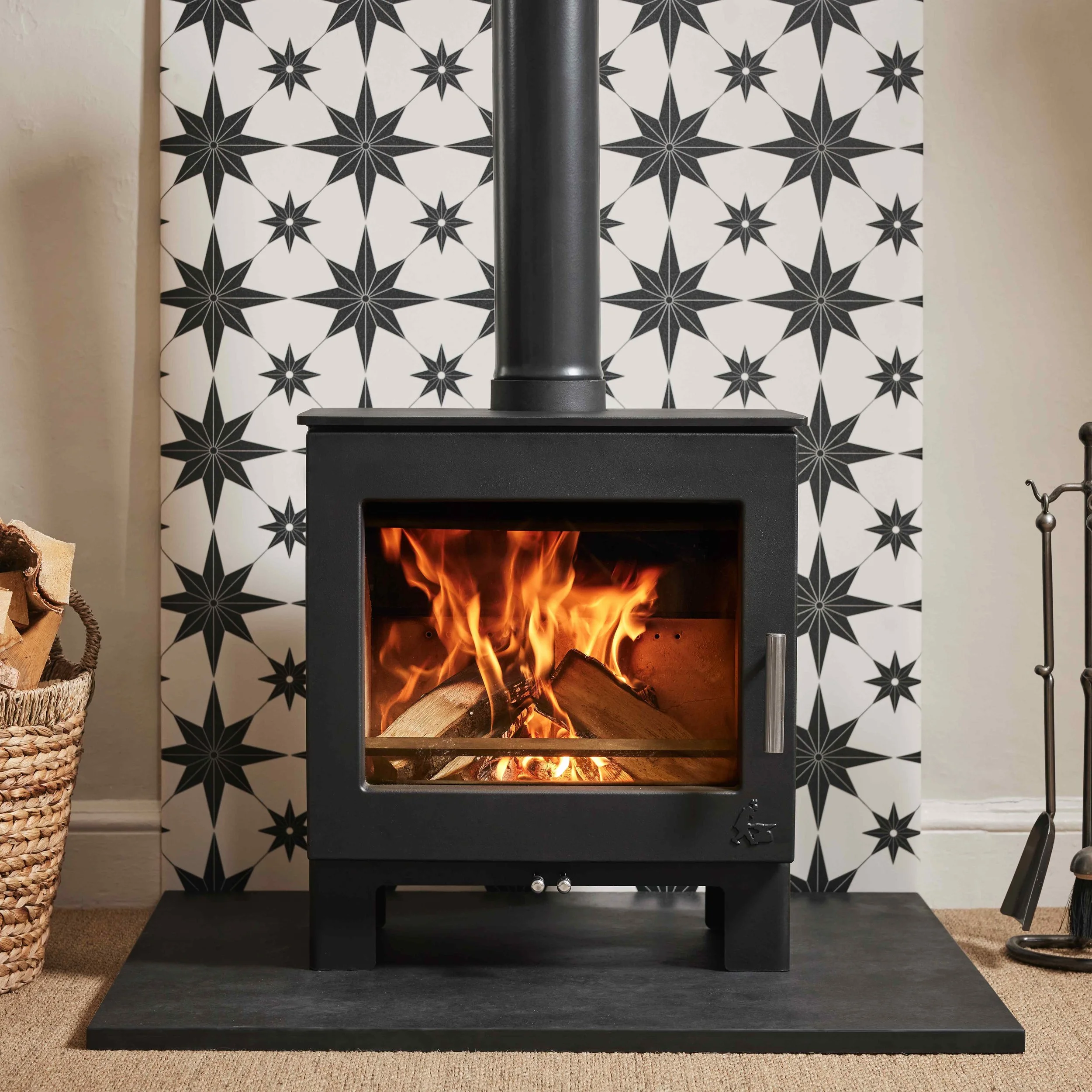 A black wood stove with glass door showing burning logs, set against a patterned black and white star design wallpaper, with a basket of firewood on the left and a metal tool on the right.
