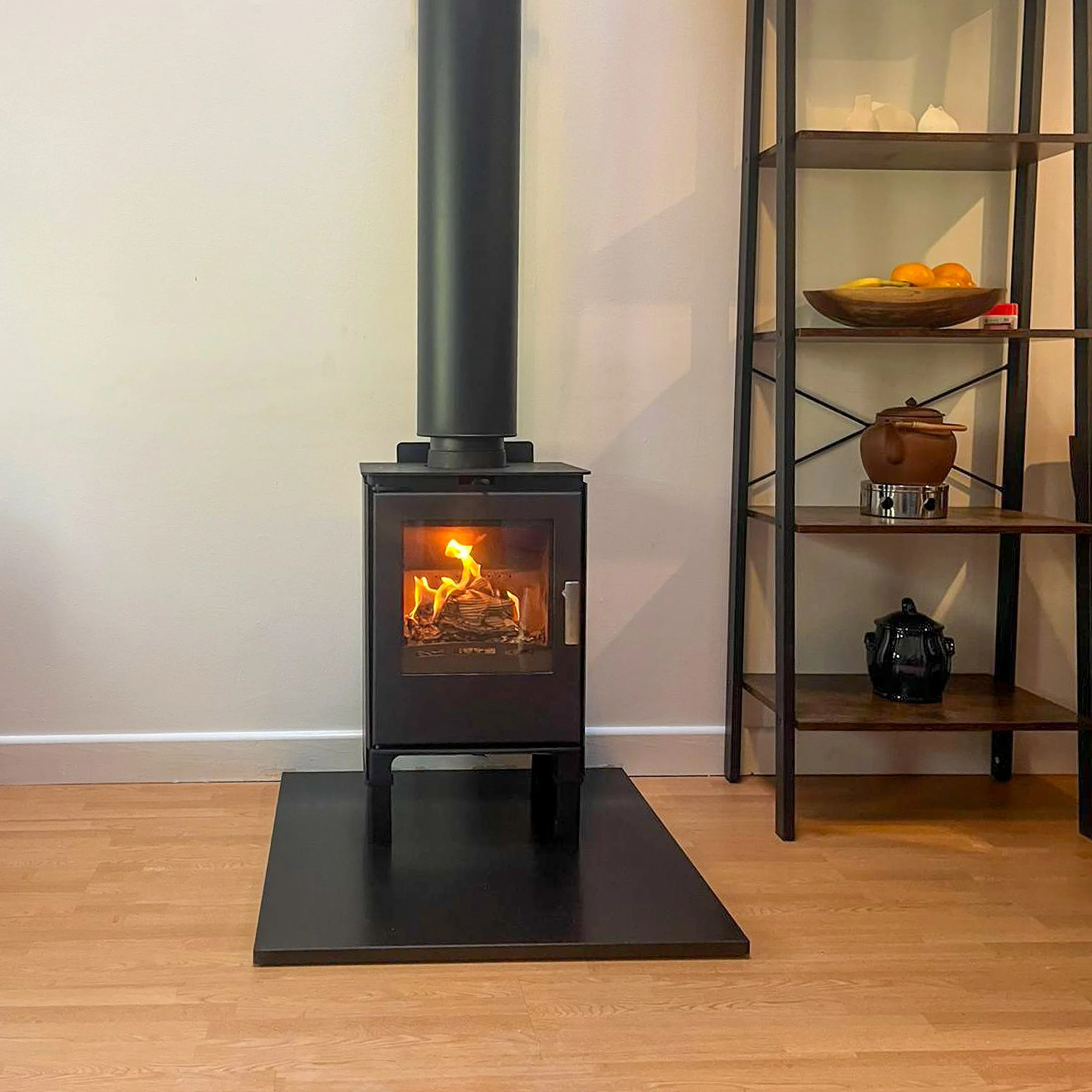 A black wood stove with a glass door showing a fire inside, standing on a black mat on a wooden floor, with a beige wall behind it. To the right, there is a black metal and wood shelving unit with decorative items including a wooden bowl of oranges, 