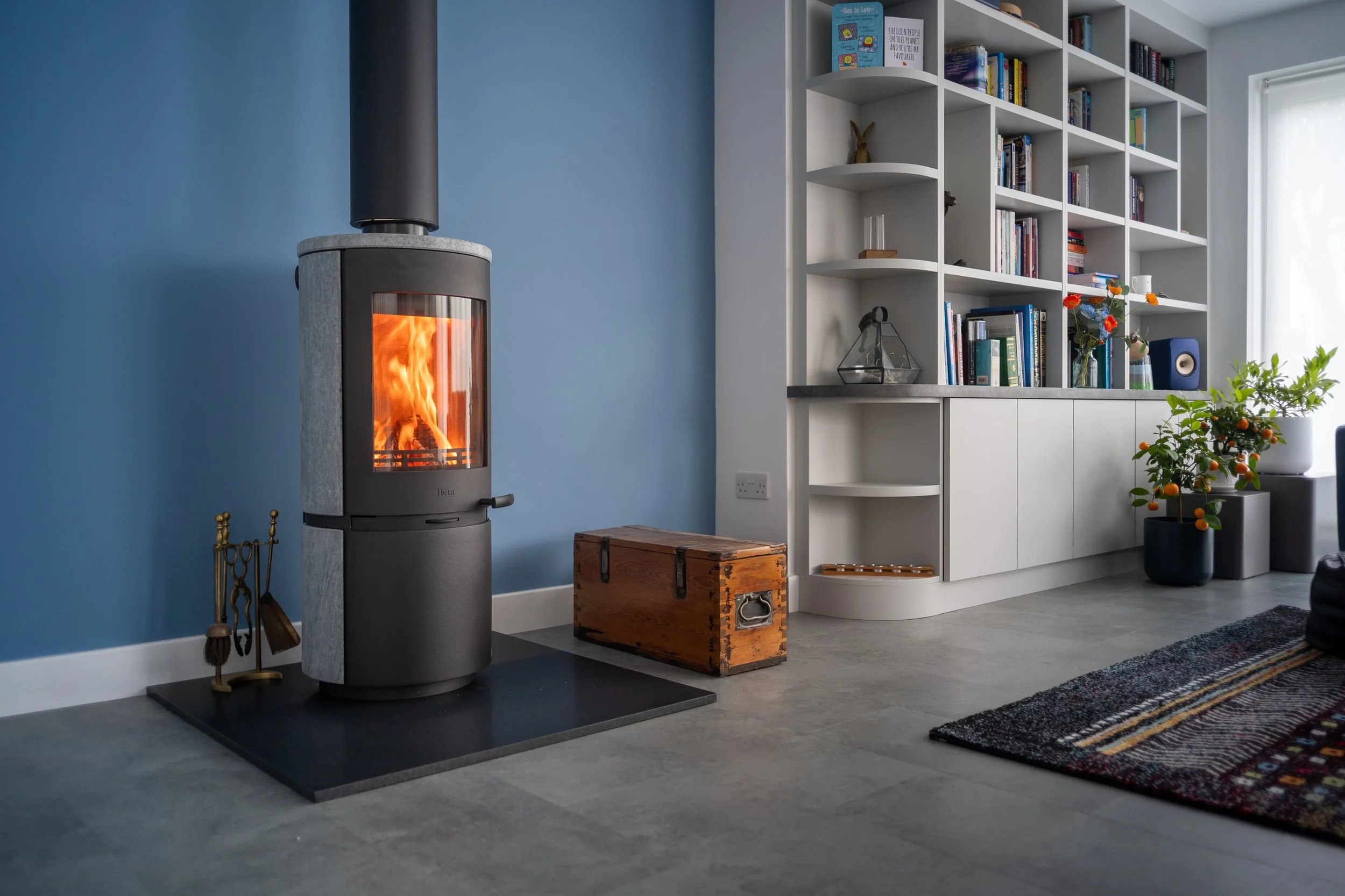 A modern living room with a wood stove emitting a visible flame, a blue accent wall, a white bookshelf filled with books and decorative items, a potted plant with orange berries, a patterned rug, and a wooden storage chest.