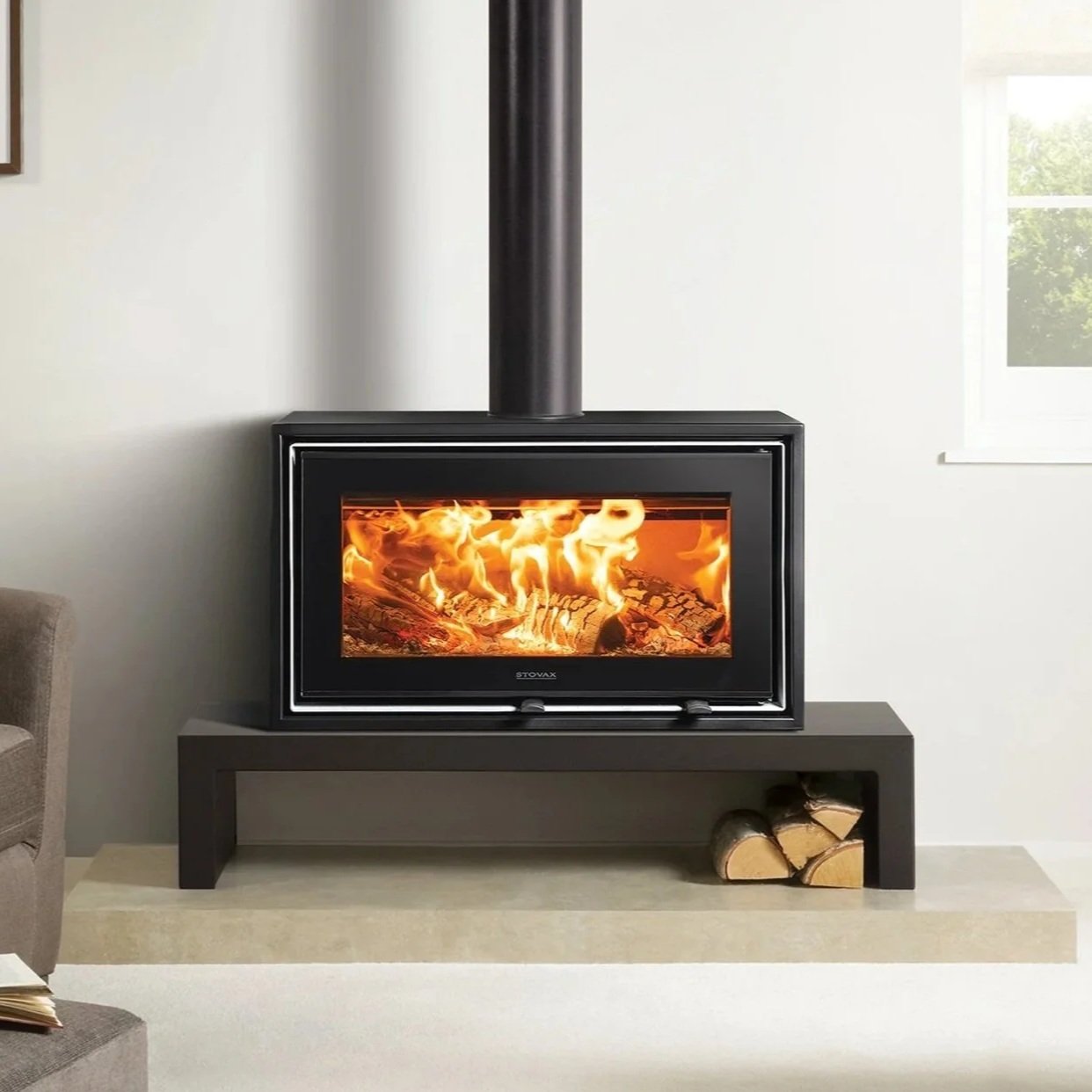 A modern black wood stove with a glass front showing burning logs inside, positioned on a black stand with firewood stored underneath, in a bright living room with a window to the right.