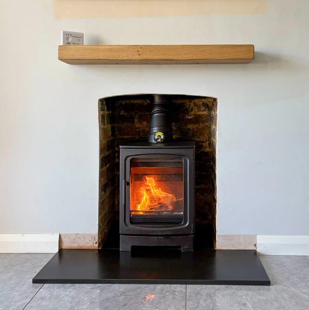 A wood stove with a fire inside, set in a brick and painted wall alcove, with a wooden shelf above and a black hearth on the tiled floor.
