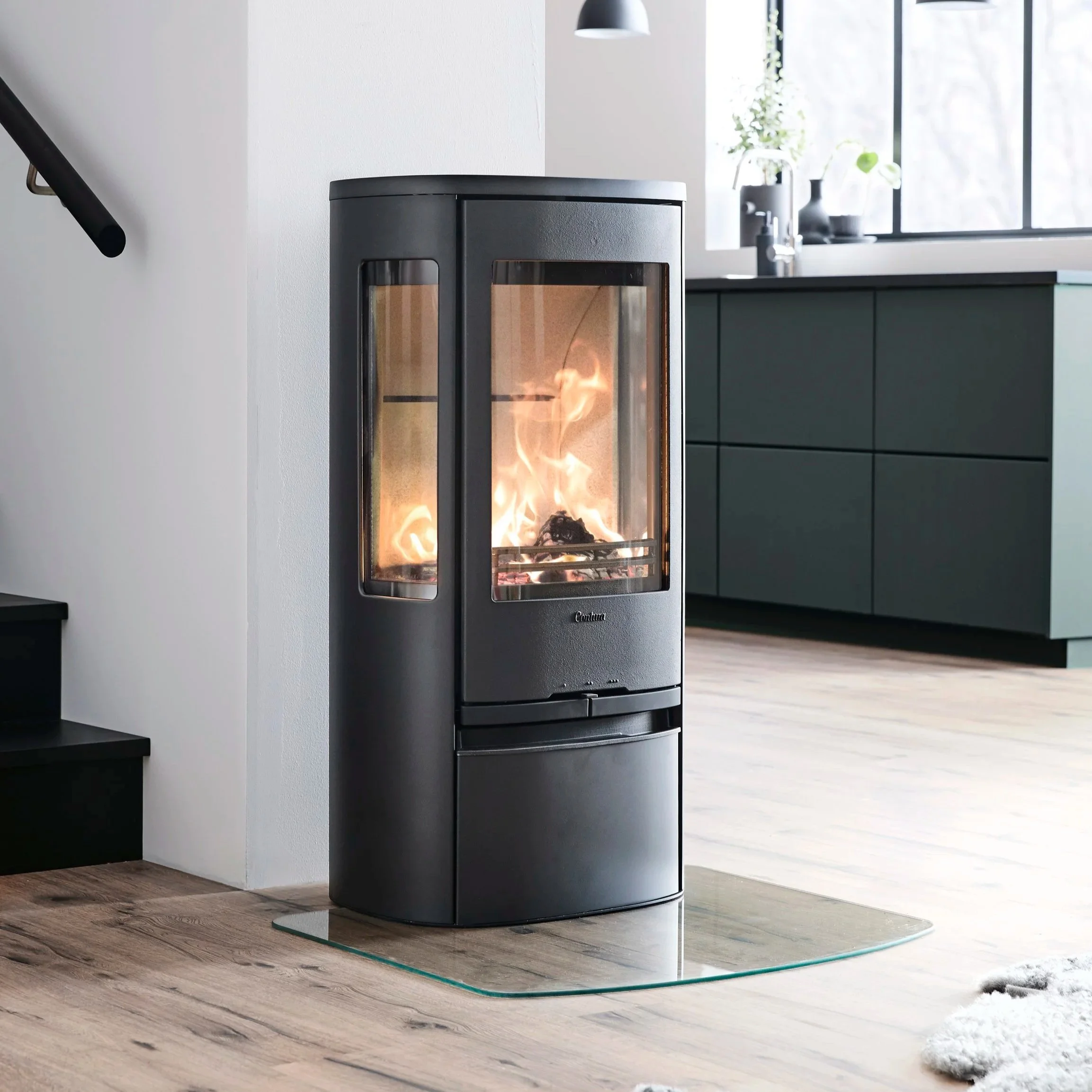 A modern wood-burning stove with a glass door showing a fire inside, situated against a white wall in a bright kitchen with large windows and dark green cabinets.