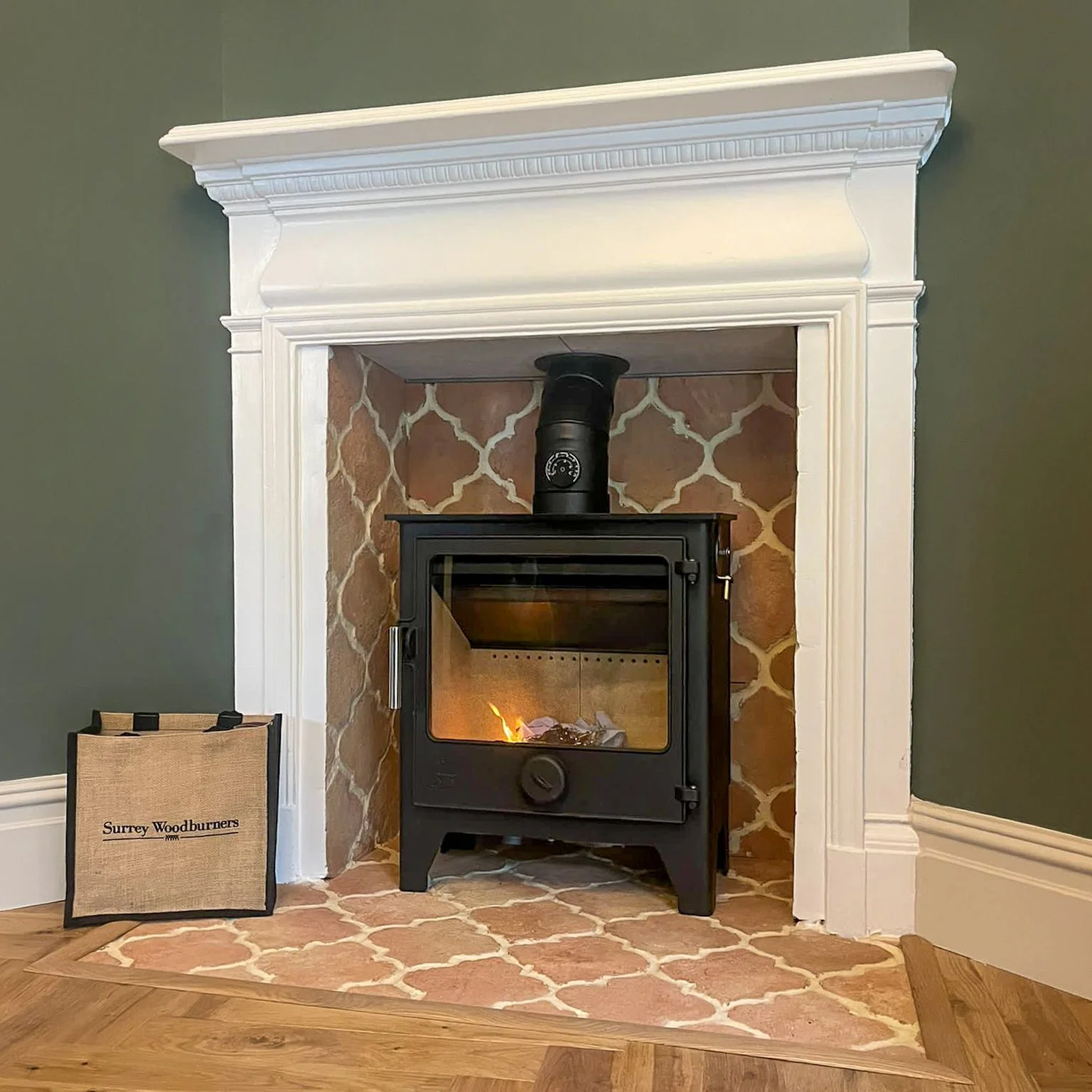 A corner fireplace with white decorative molding, a black wood stove inside, and a bag labeled Surrey Woodburners next to it.