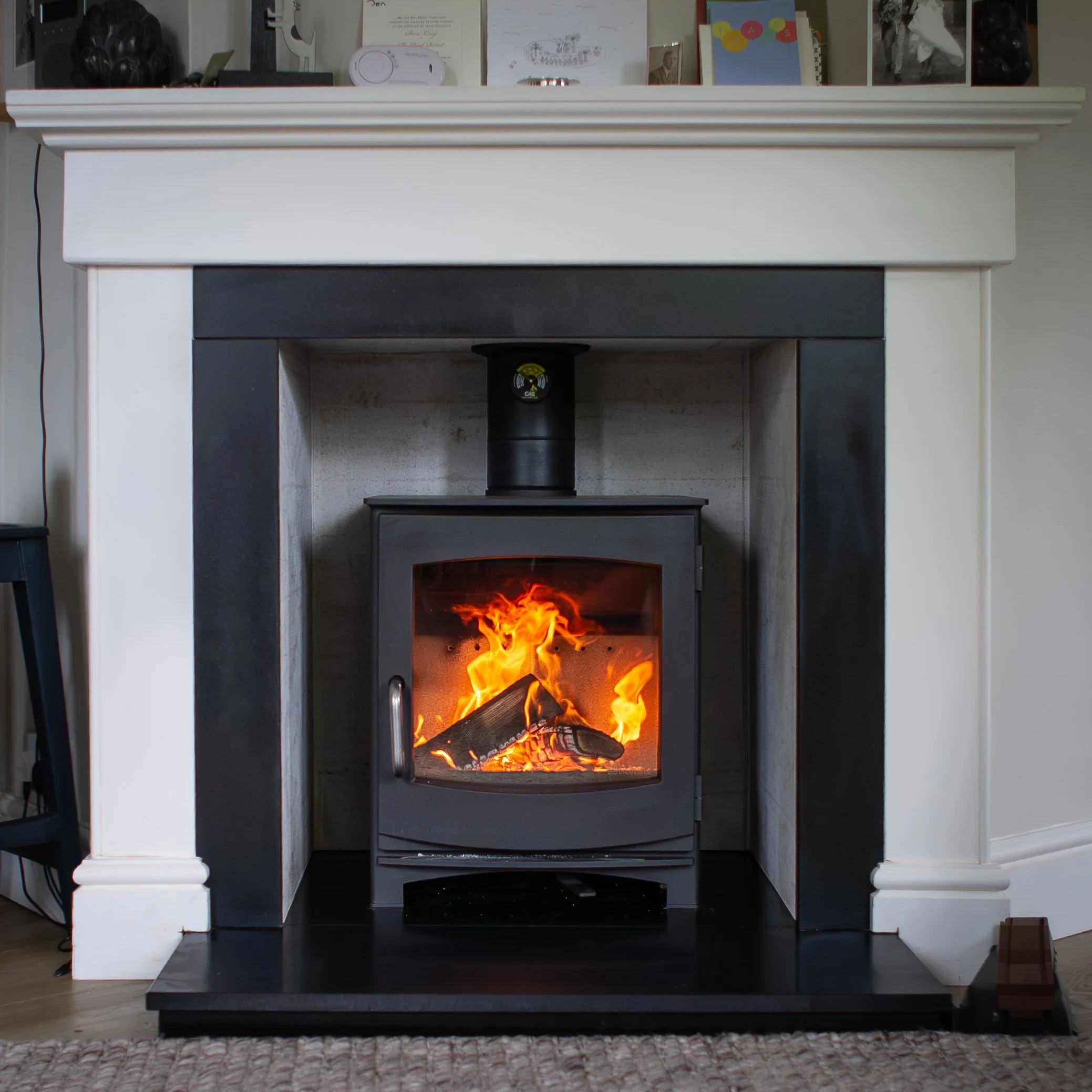 A black fireplace with a wood-burning stove inside, flames visible through a glass door, and a white mantel above with family photos and decorative items.