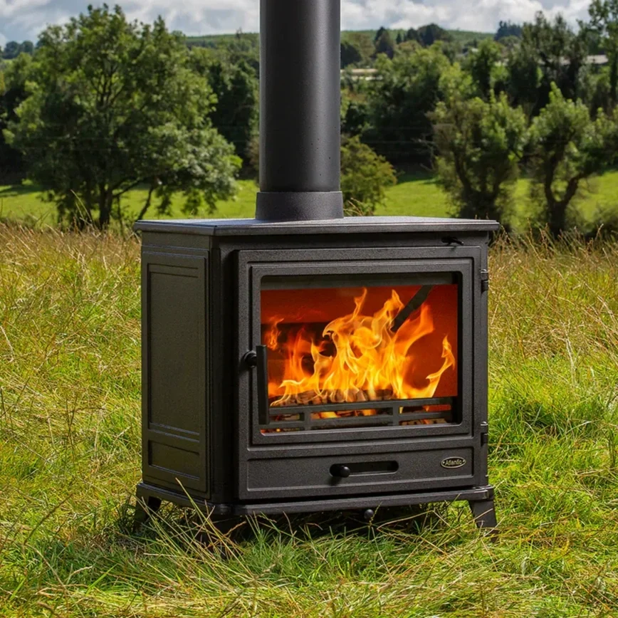 A black wood-burning stove with a glass door showing orange flames inside, situated outdoors on a grassy field with trees and a cloudy sky in the background.