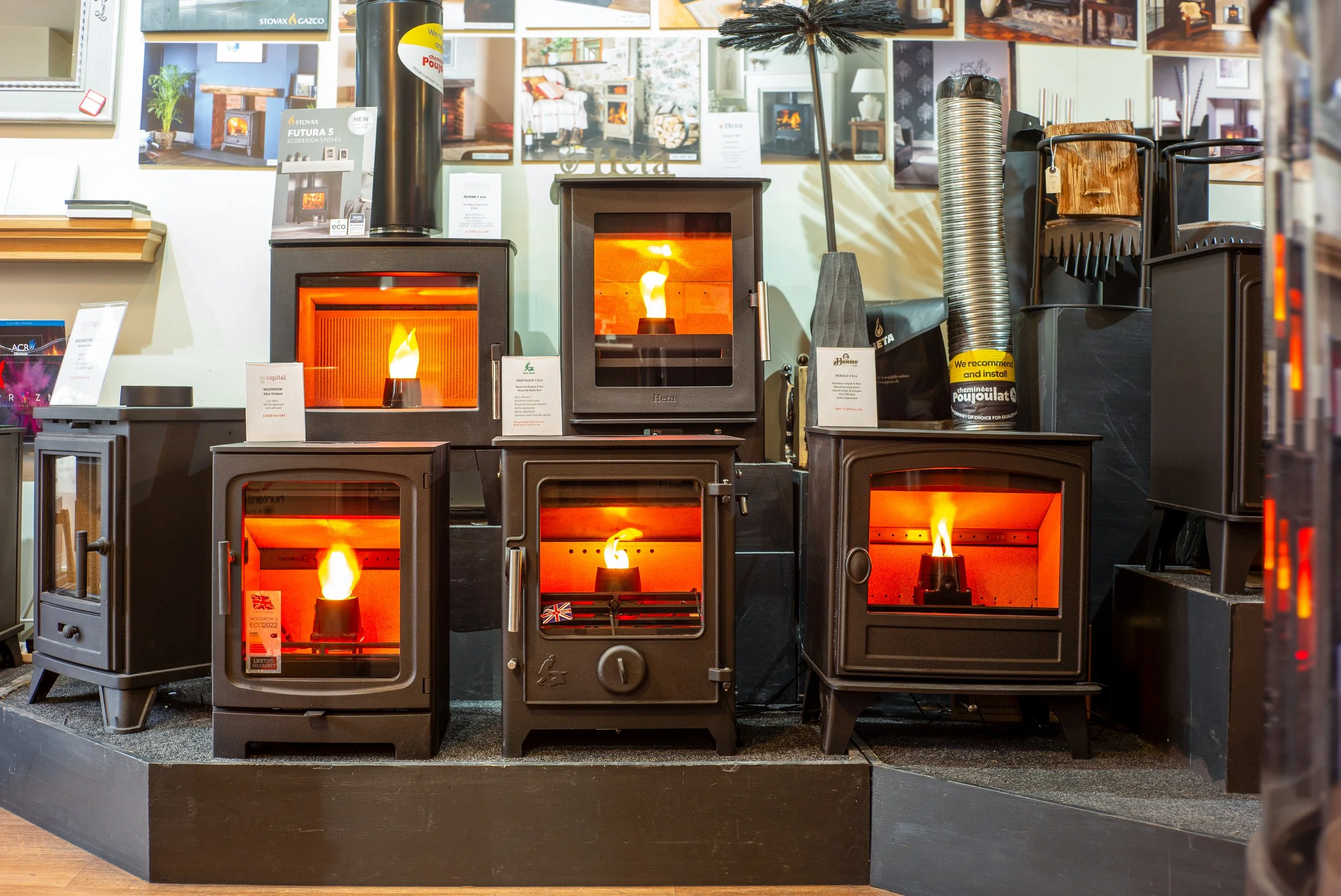 Display of various wood-burning stoves in a showroom, showcasing different models with glass doors and flames inside.