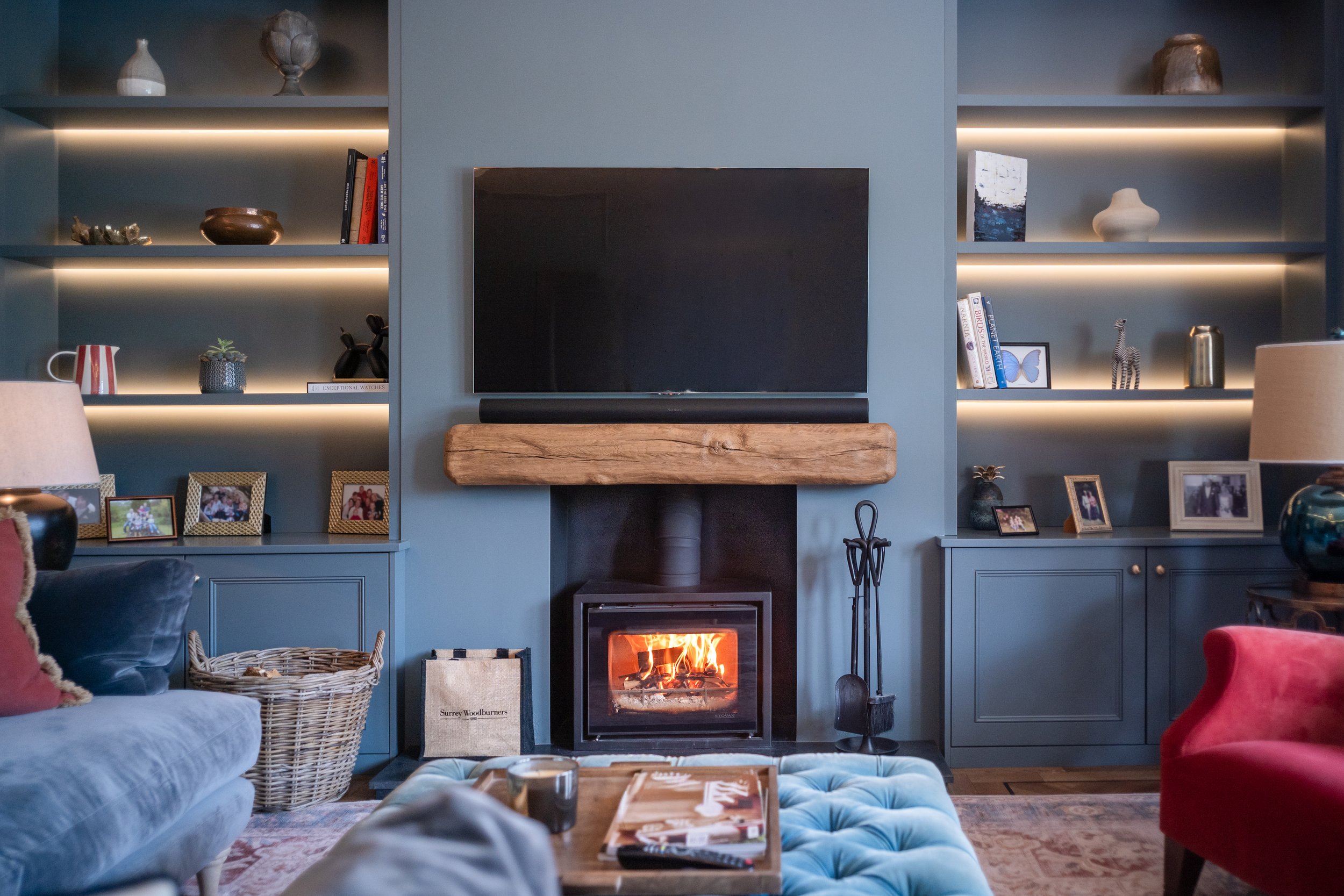 Living room with a blue built-in bookshelf, a wall-mounted TV, and a fireplace with a fire burning. The shelves hold decorative items, books, and framed photos. There is a couch with pillows, a basket, and a coffee table with magazines and candles.