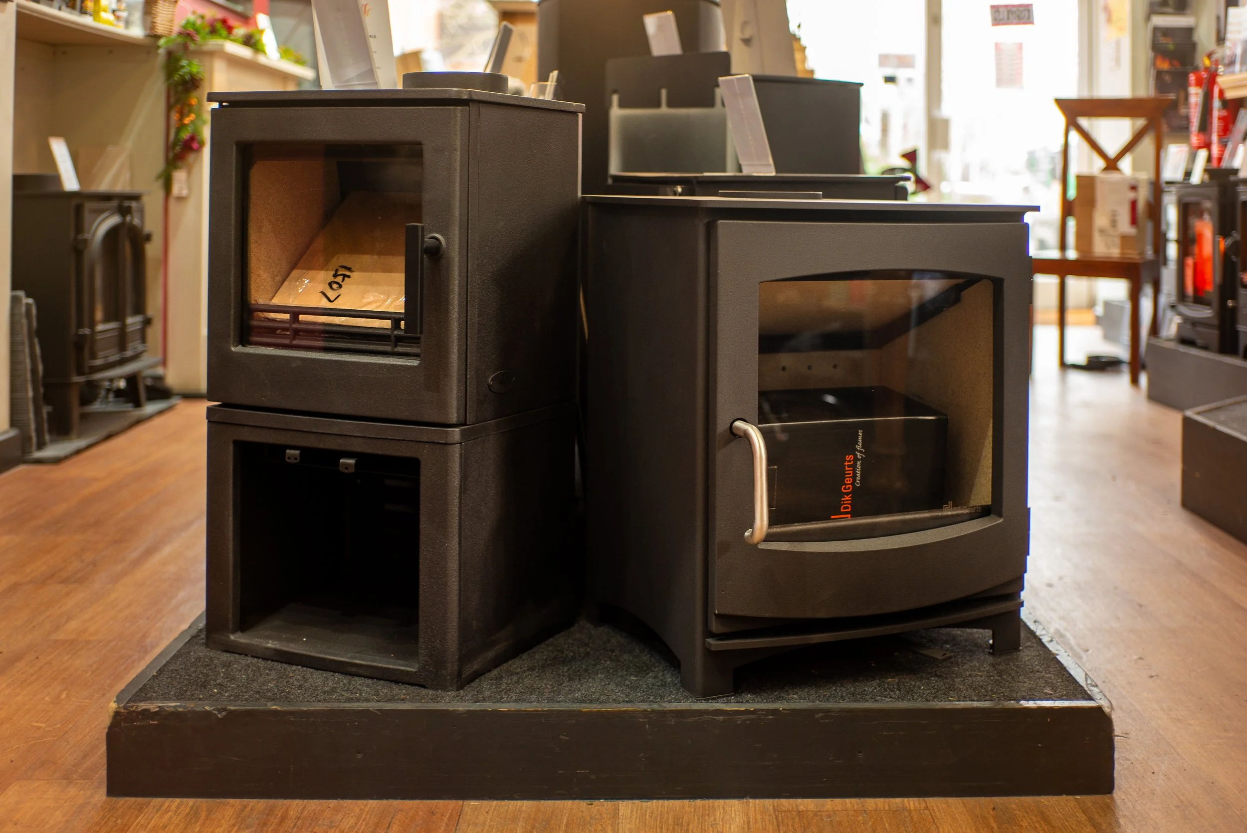 Three black wood stoves or fireplaces displayed on a wooden floor inside a store, with various other home decor items visible in the background.