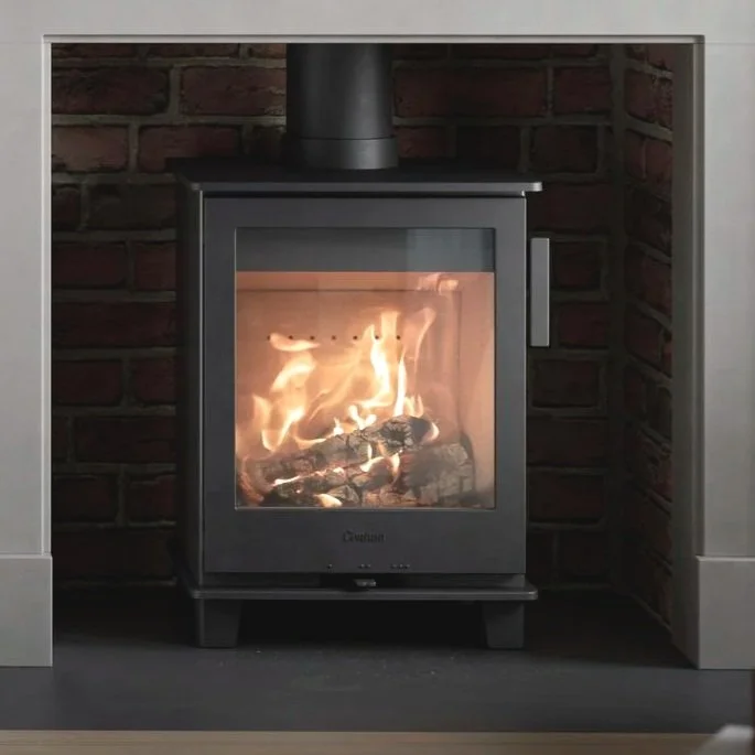 A modern fireplace with a glass front showing a burning fire with logs inside, set against a brick wall.