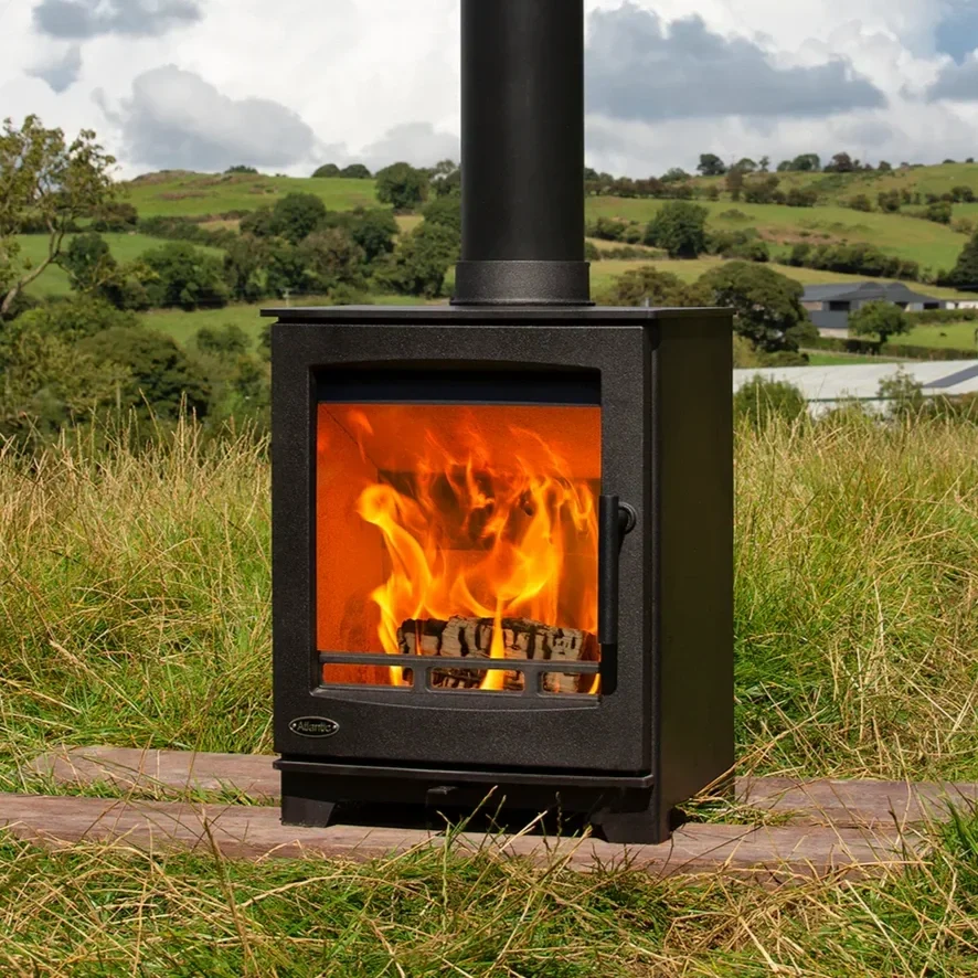 A black wood stove with a glass door showing a fire inside, situated outdoors on a grassy field with rolling hills and trees in the background.