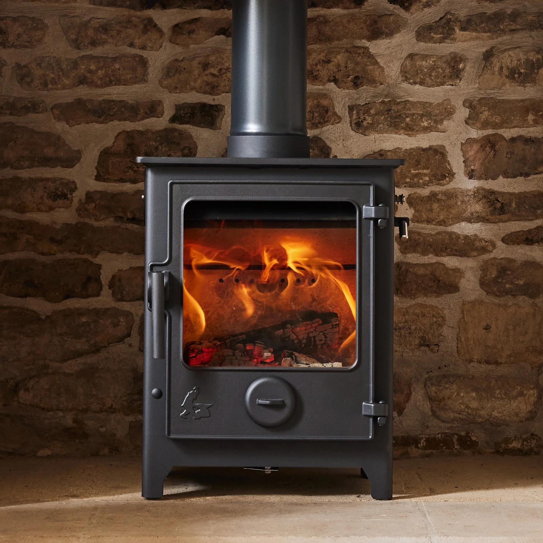A black wood stove with visible fire inside, set against a rustic brick wall.