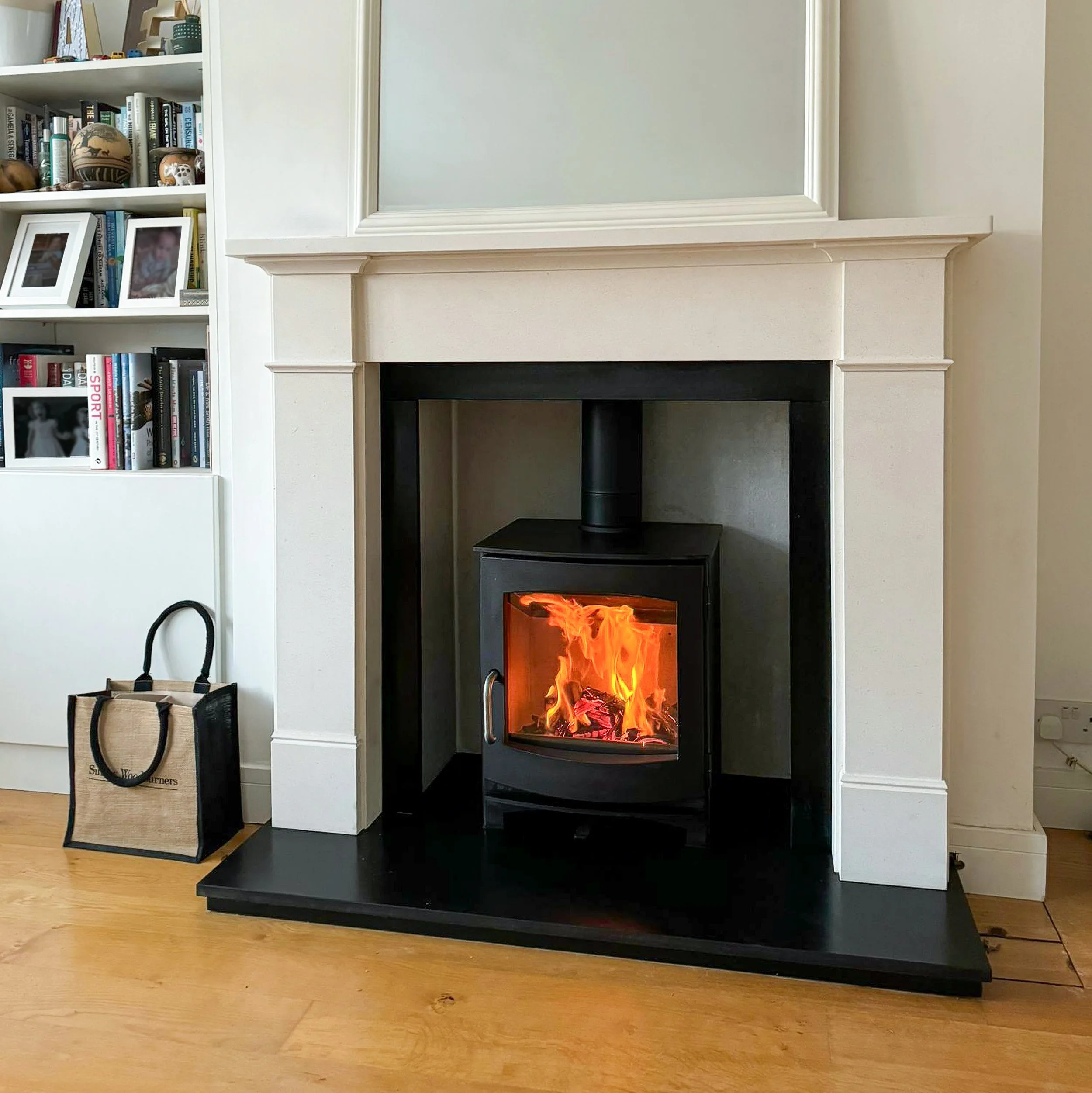 A cozy living room fireplace with a roaring fire in a black wood stove, surrounded by a white mantel and black hearth, with a bookshelf and decorative items in the background.