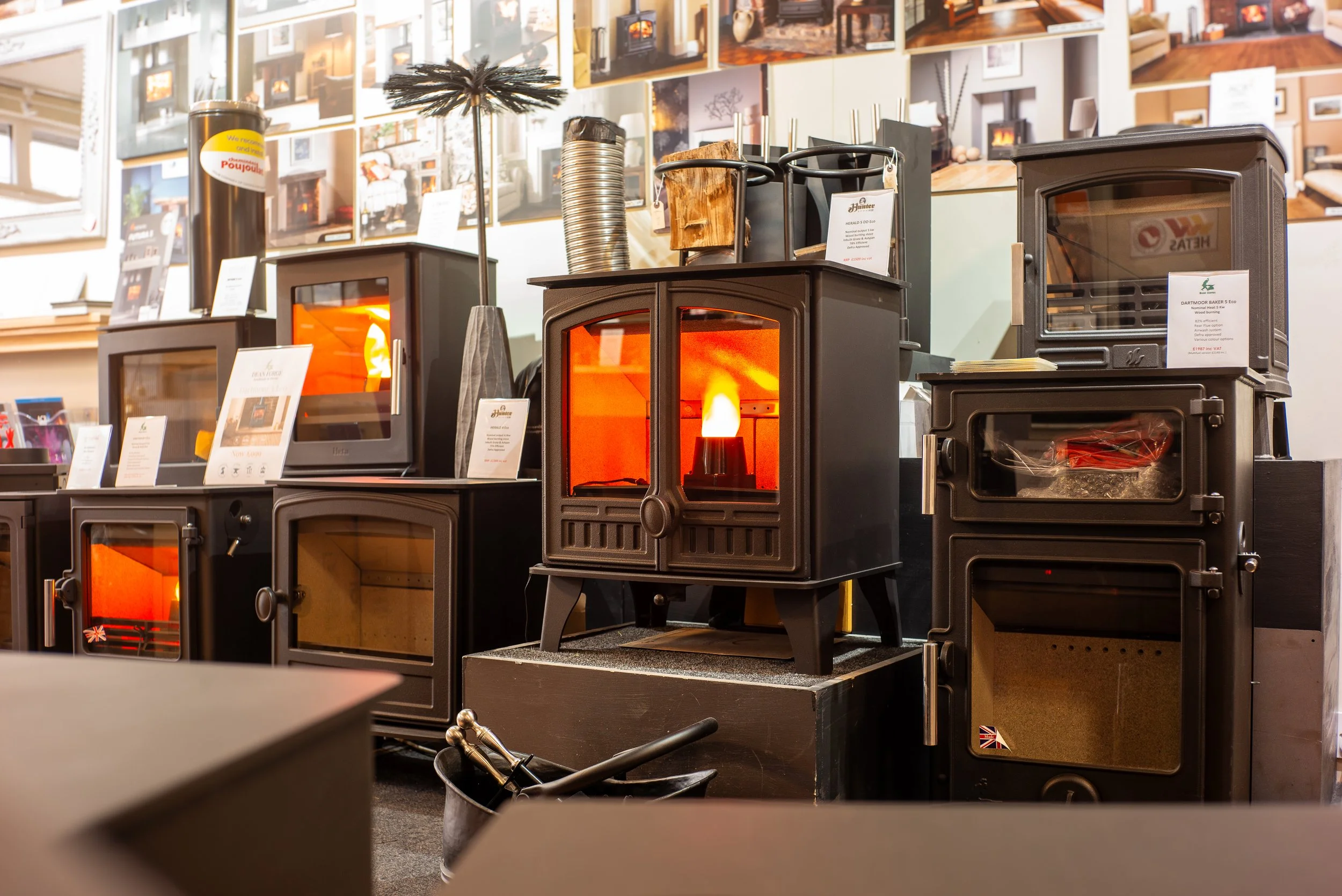 Display of various wood-burning stoves and fireplaces in a showroom, with framed pictures on the wall behind.