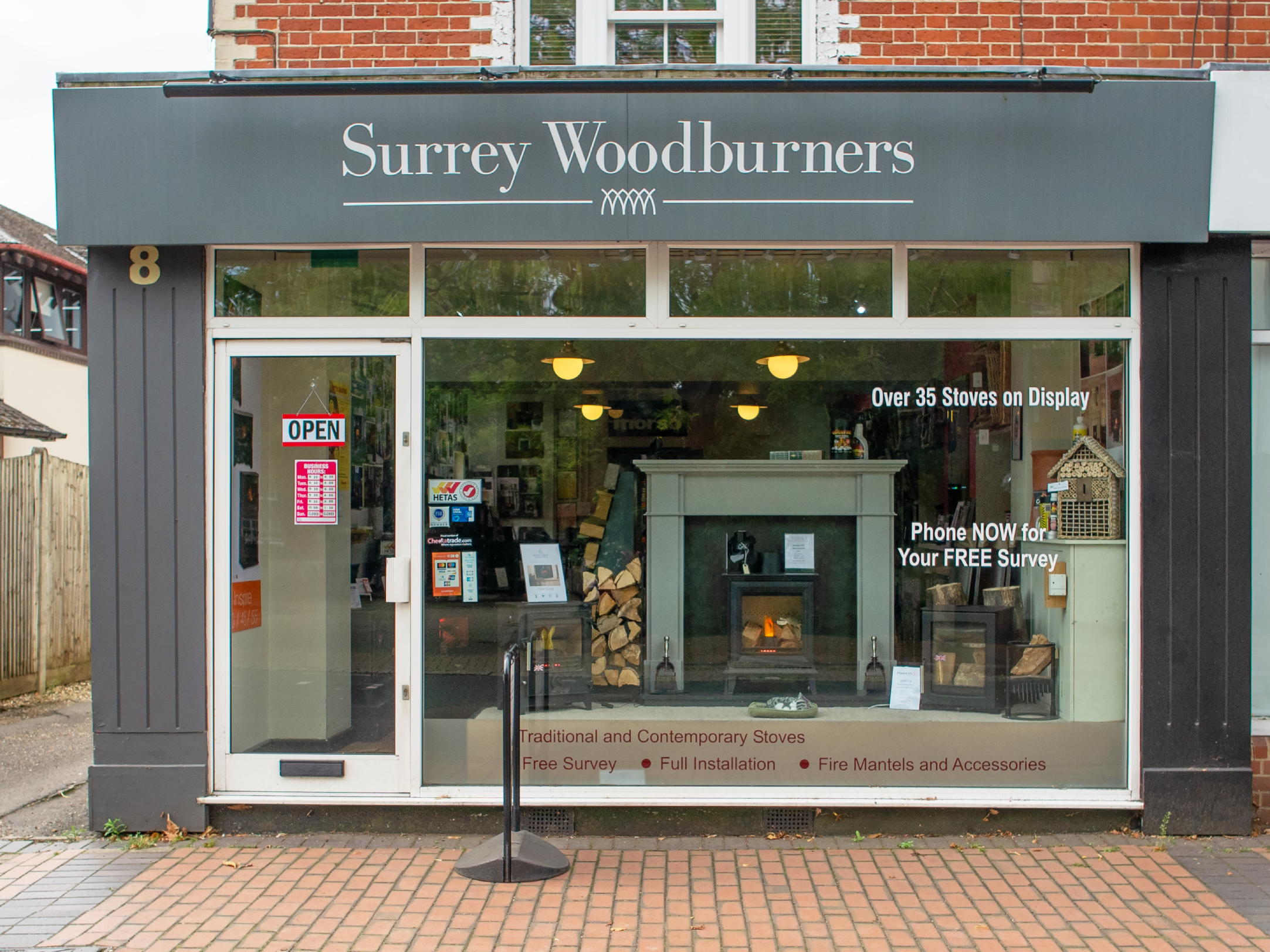 storefront of Surrey Woodburners with a black sign and white text, glass window showing fireplaces and firewood inside, and an open sign on the door. The storefront advertises over 35 stoves on display, free survey, full installation, and fire mantels and accessories.