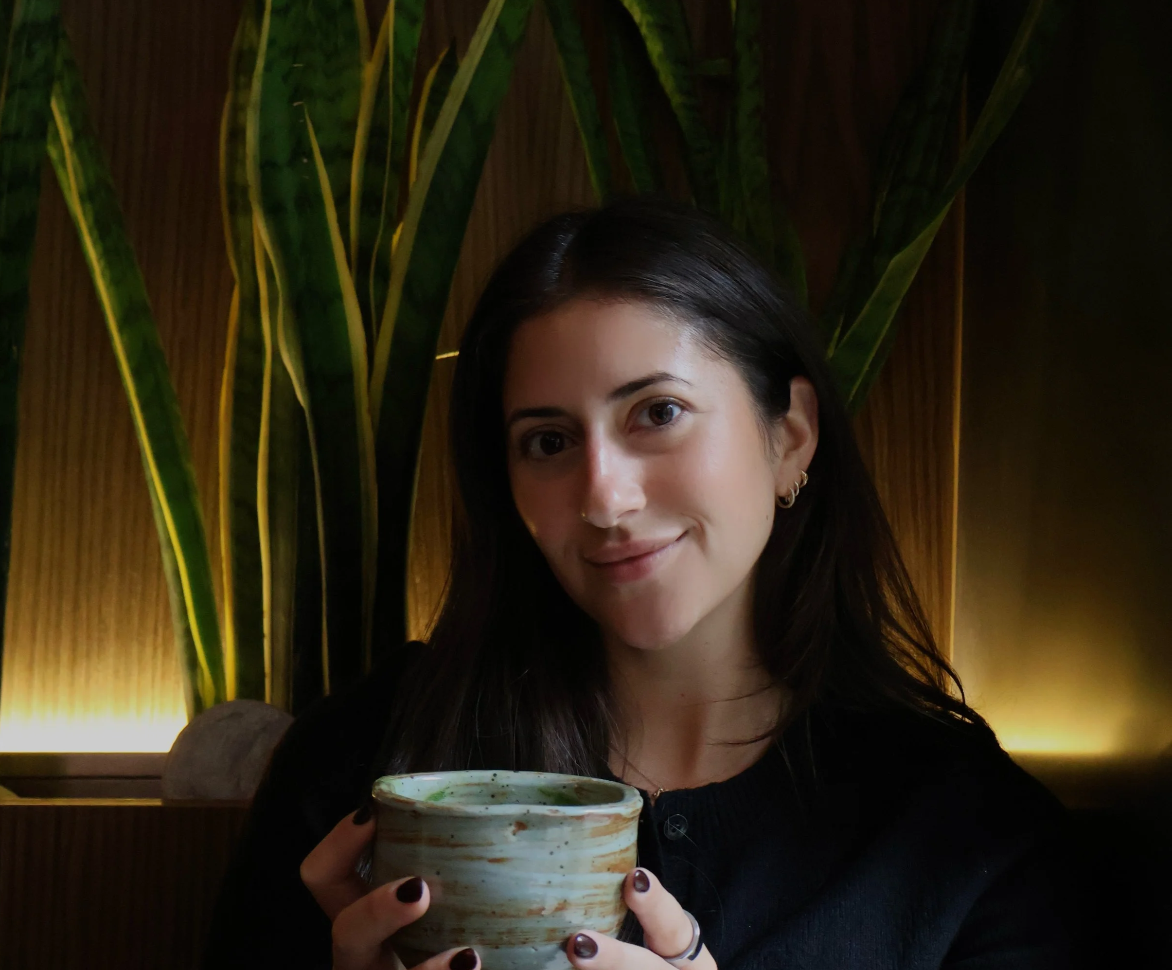 A woman with dark hair and earrings holding a ceramic bowl and smiling indoors with green plants and warm lighting in the background.