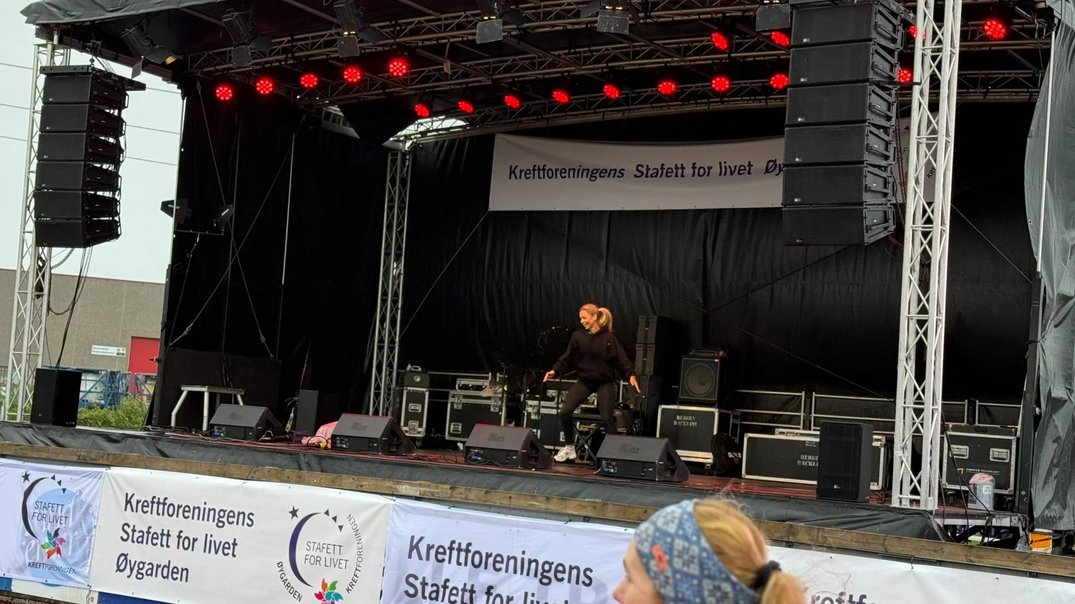 En person opptrer på en utendørs scene med lydutstyr, med et banner som sier "Kreftforeningens Stafett for livet Øygarden".