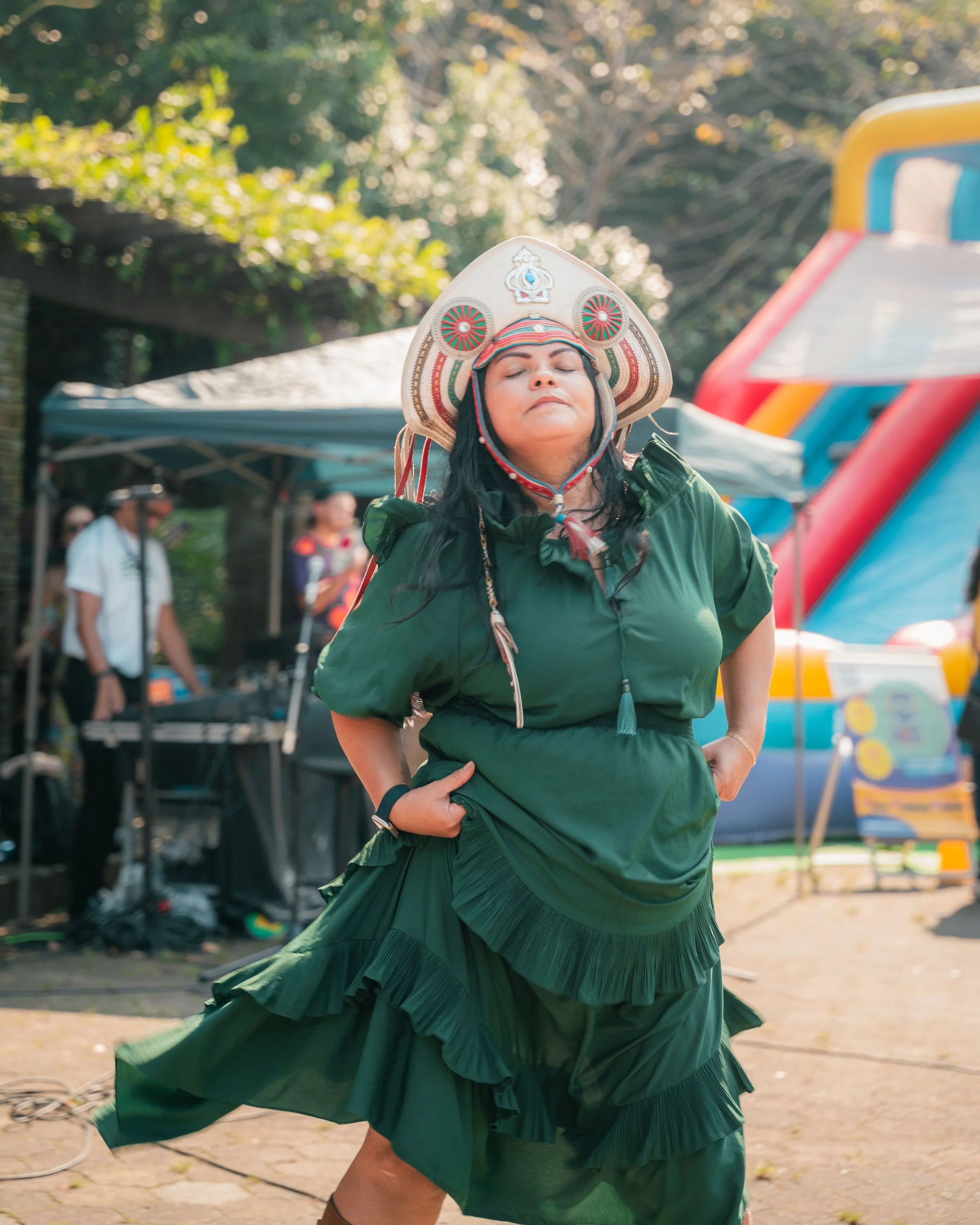 A woman performing a traditional indigenous dance outdoors, wearing a green dress with ruffles and an elaborate embroidered hat with red, white, and blue details. In the background, there are tents, people, and an inflatable slide.