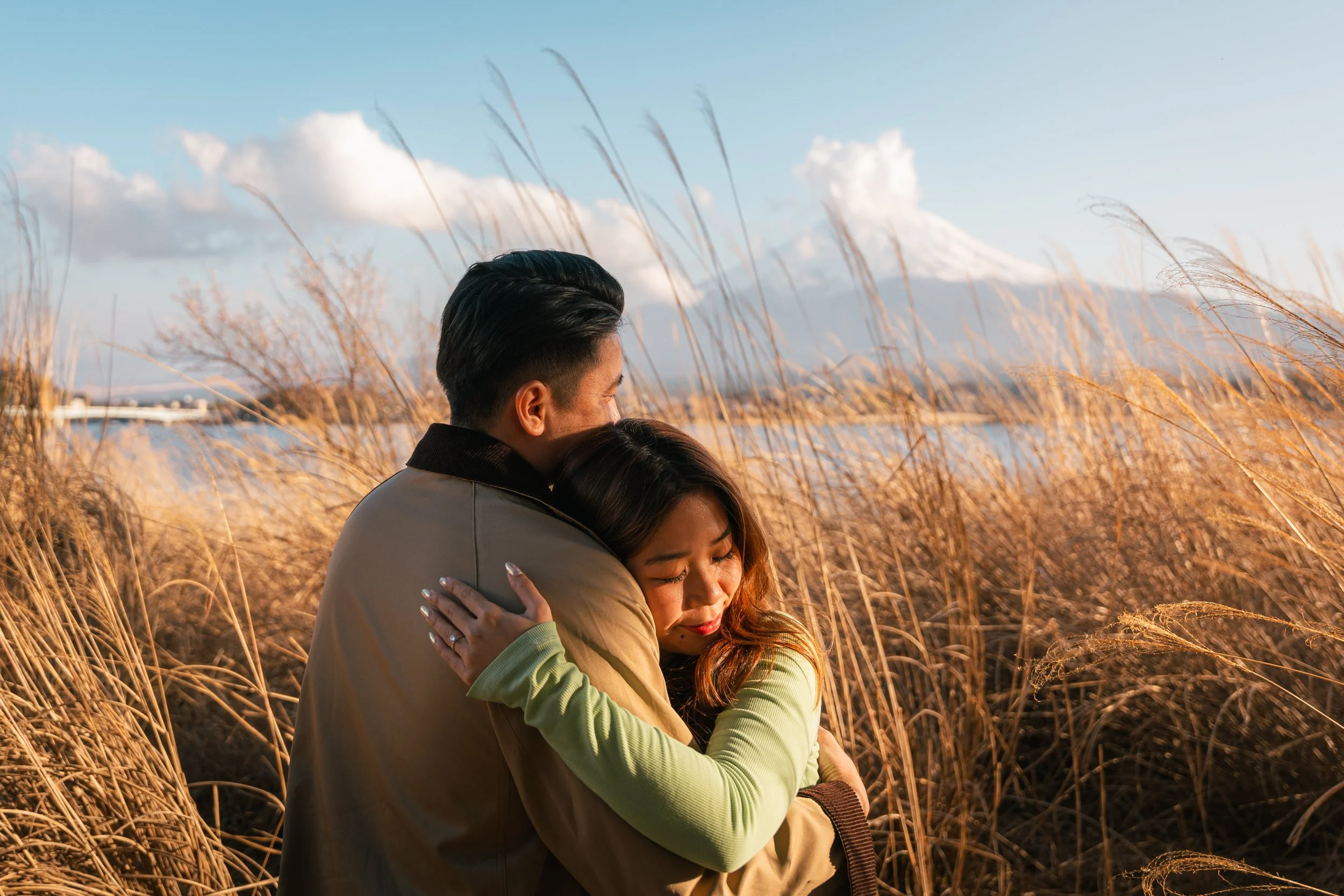 A man and woman embracing in a field of tall, golden grasses with a mountain and blue sky in the background