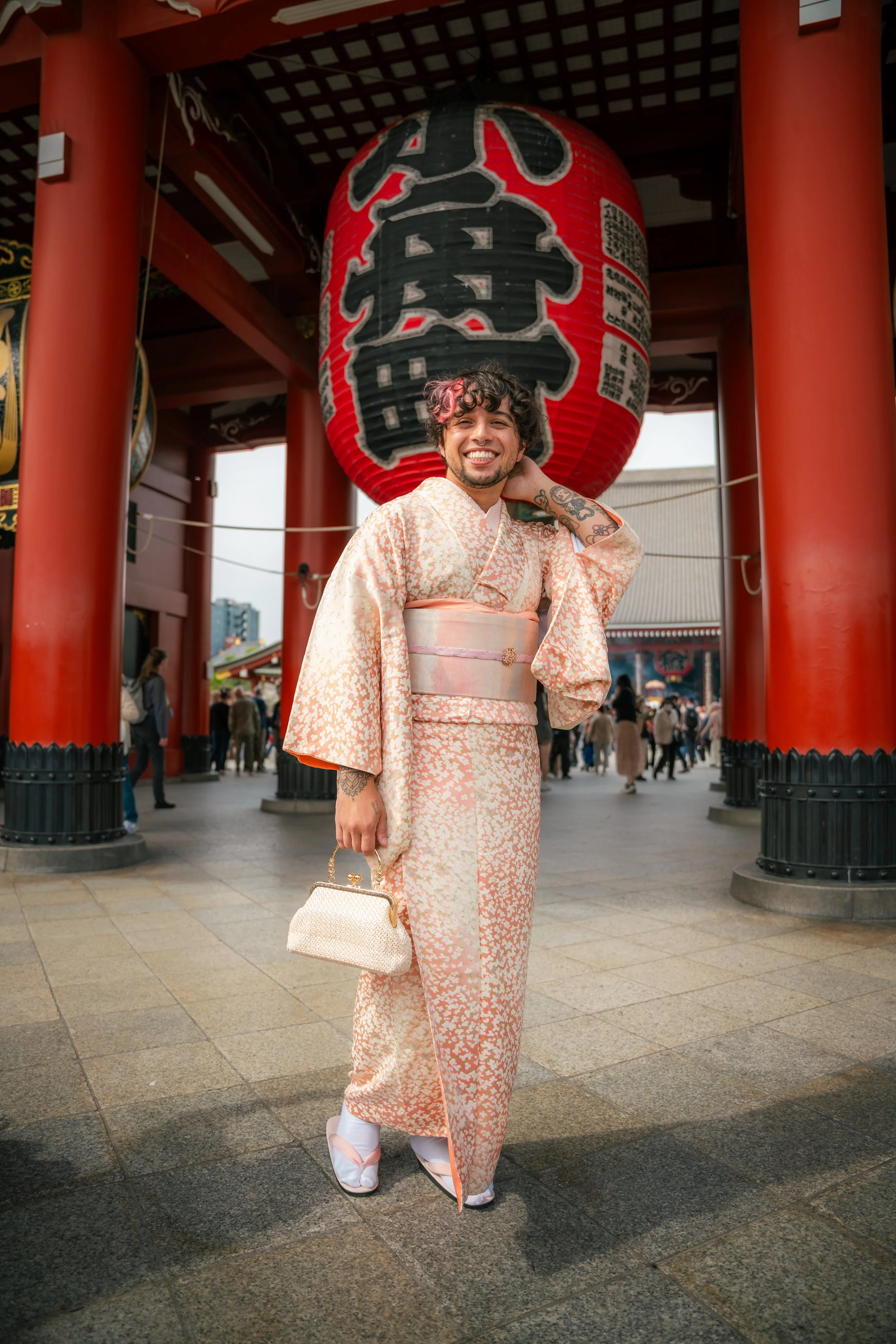 A smiling man dressed in a light-colored kimono with a pink and white pattern, holding a small beige handbag, standing in front of a traditional Japanese temple gate with a large red paper lantern.