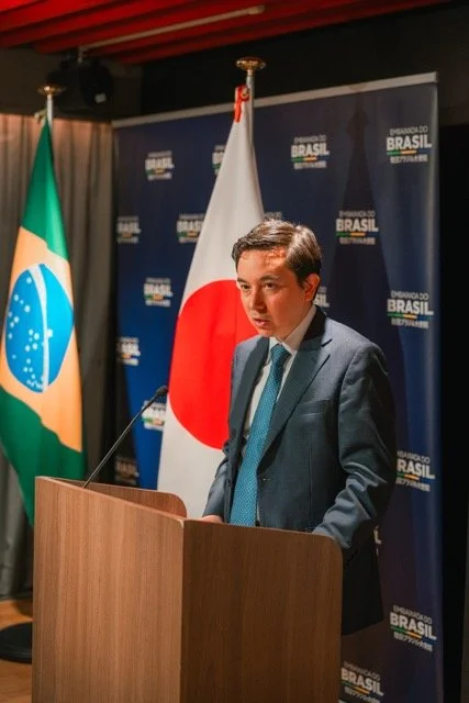 A man in a suit standing at a podium with a microphone, with the flags of Brazil and Japan behind him during a press conference or official event.