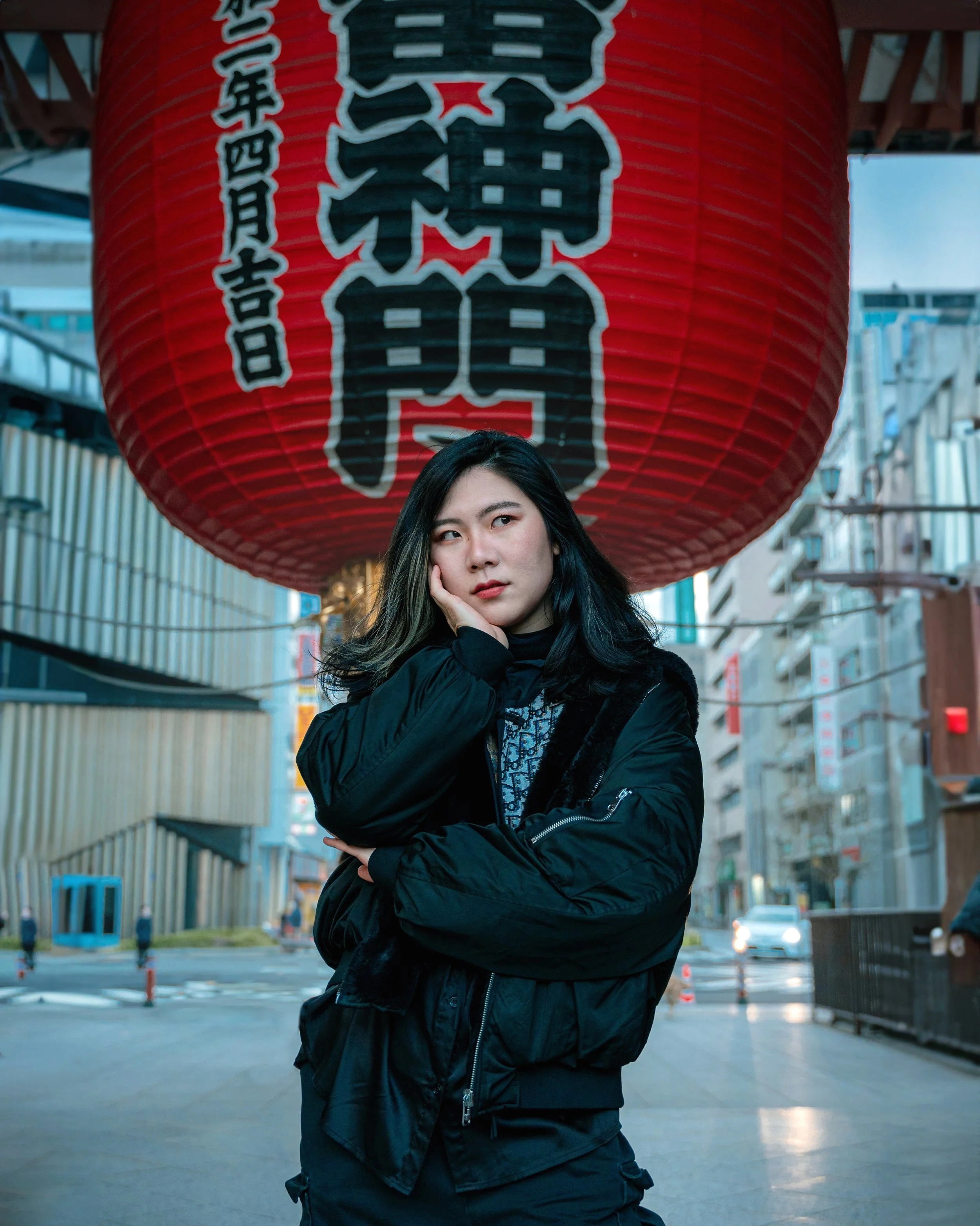 A young woman stands in front of a large red Japanese paper lantern with black and white characters, wearing a black jacket, on an urban street.