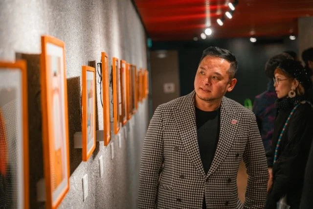 A man in a checkered blazer looking at framed artwork on a gallery wall with other people in the background.