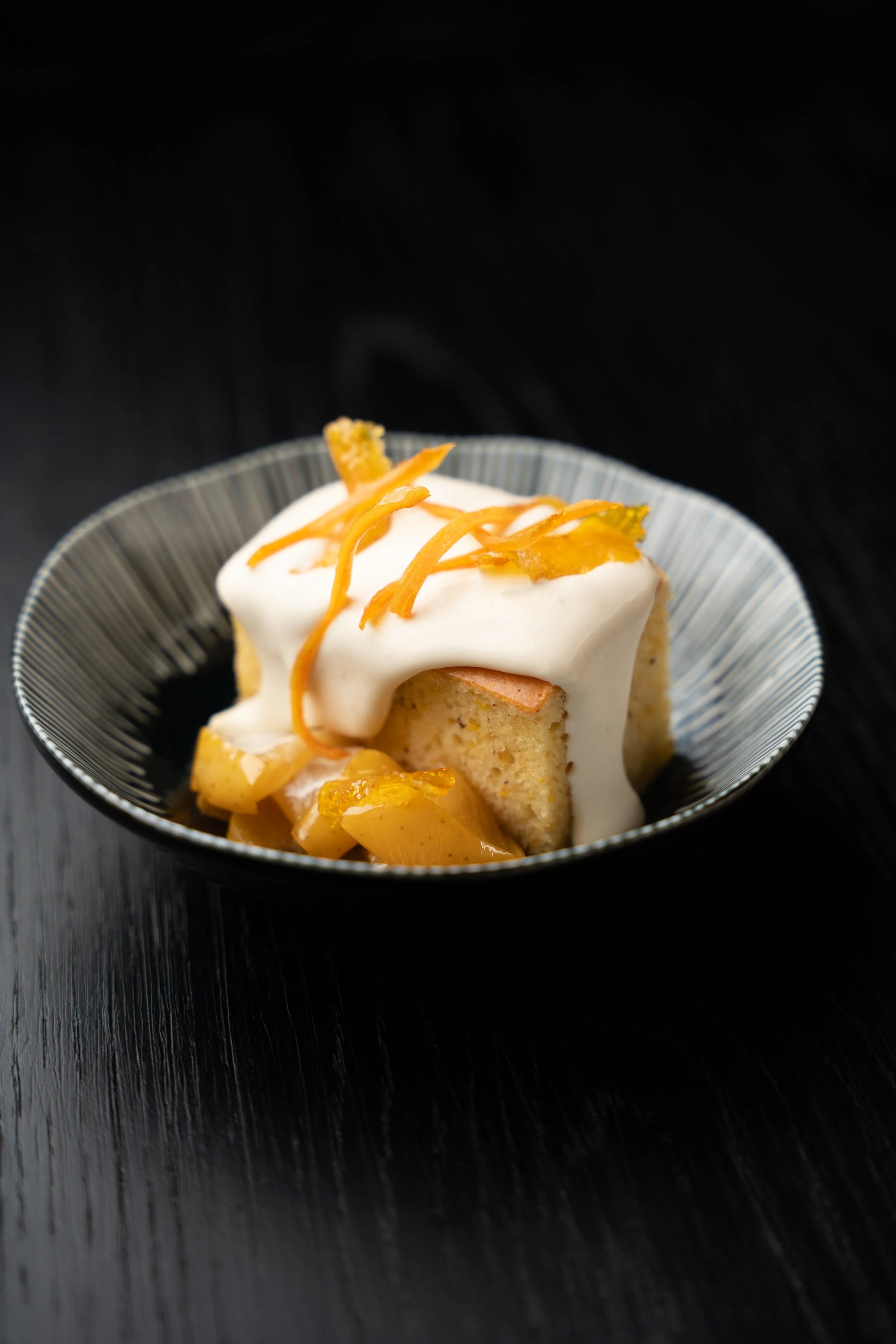 A square piece of cake with white icing and yellow decorative shreds on top, served in a striped black and white bowl on a black surface.