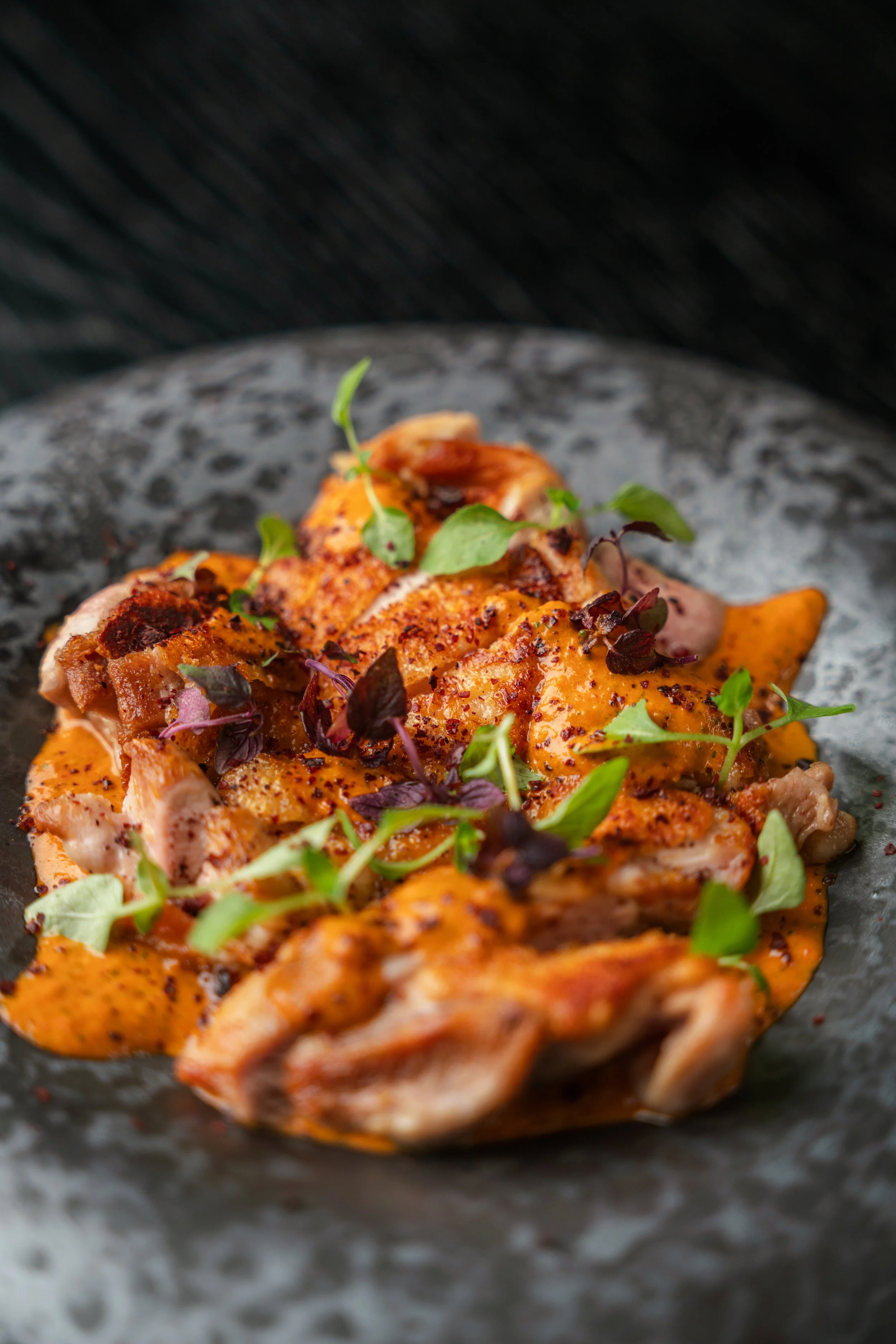 Close-up of grilled chicken pieces topped with sauce and microgreens on a dark plate.