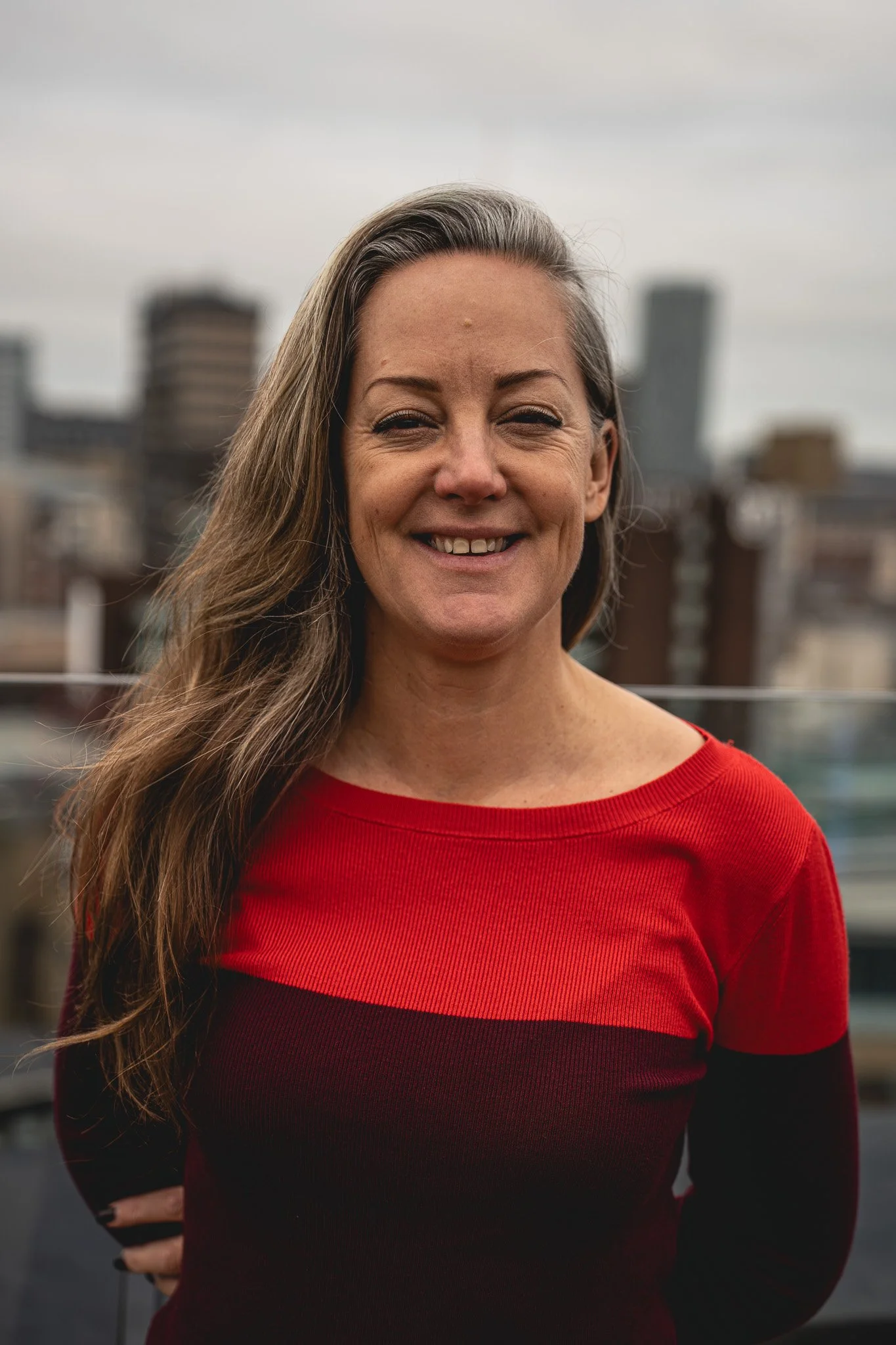 A smiling Zandra Moore MBE, CEO and Founder of Zygens, with long brown hair in a red and black sweater standing outdoors with Leeds city buildings in the background.