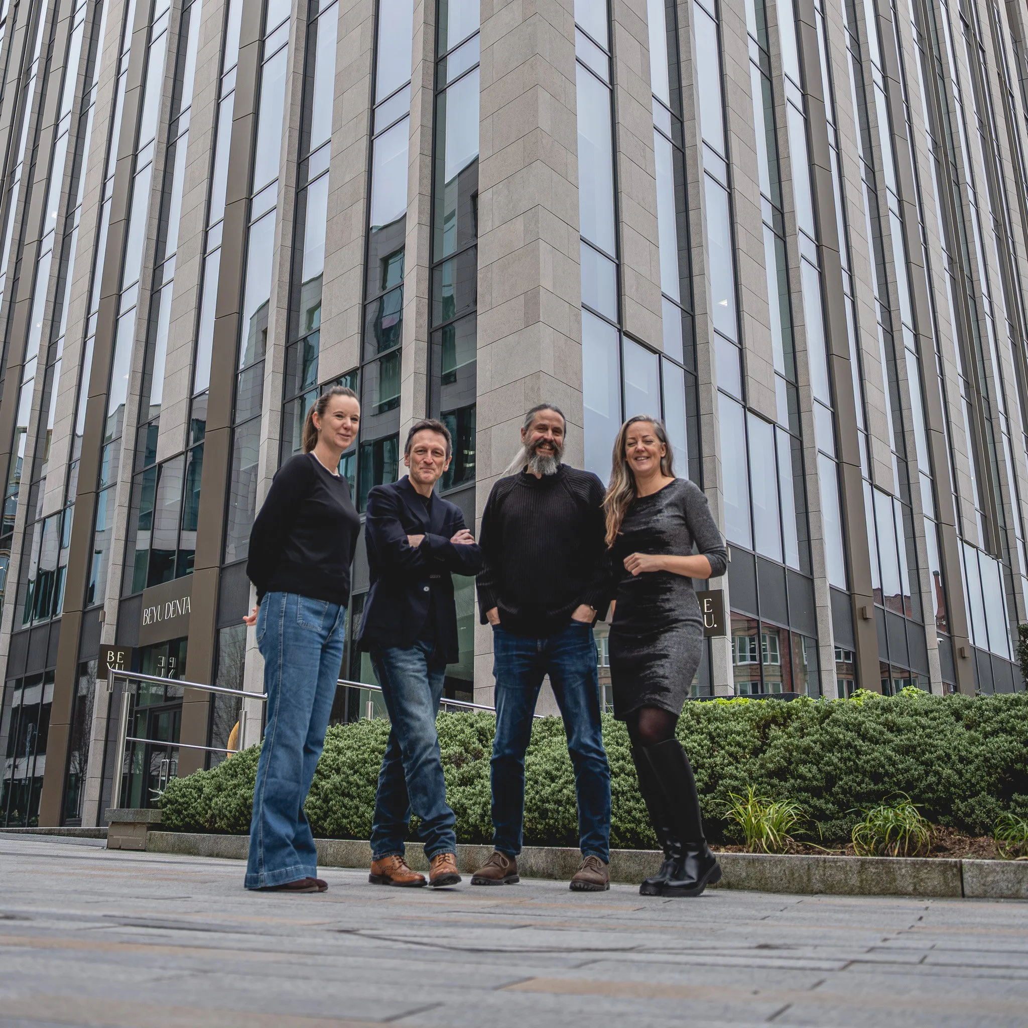 The four Zygens founders - L-R Charlie Bartle, Andy Roberts, Tim Lewis and Zandra Moore MBE people standing outside in front of a modern office building in Leeds, smiling and posing for a photo.