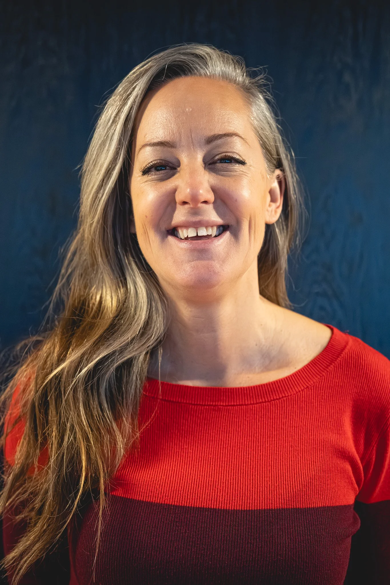 A woman with long, wavy blonde hair smiling, wearing a red top with black horizontal stripes against a dark blue background.