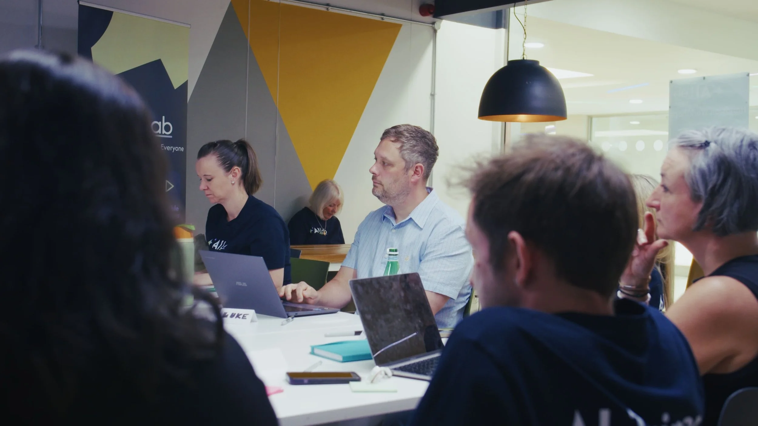 People attending a meeting or conference in a modern office space, working on laptops and listening attentively, with a focus on the individual in the center wearing a light blue shirt.