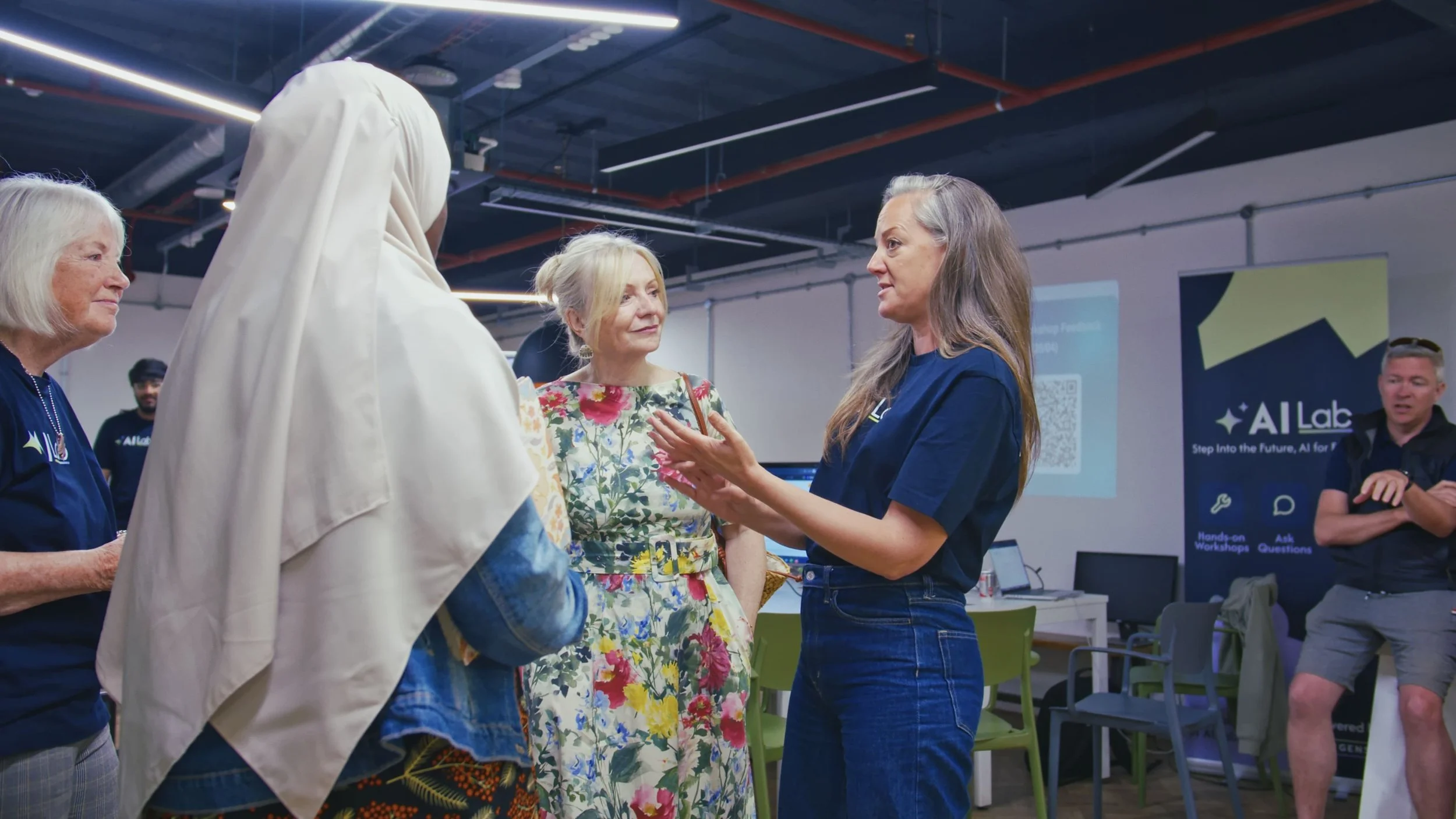 A group of women, including a woman in a floral dress and a woman with long gray hair, are engaged in conversation with a woman in a blue shirt at an indoor event. There are informational signs and a presentation screen in the background.