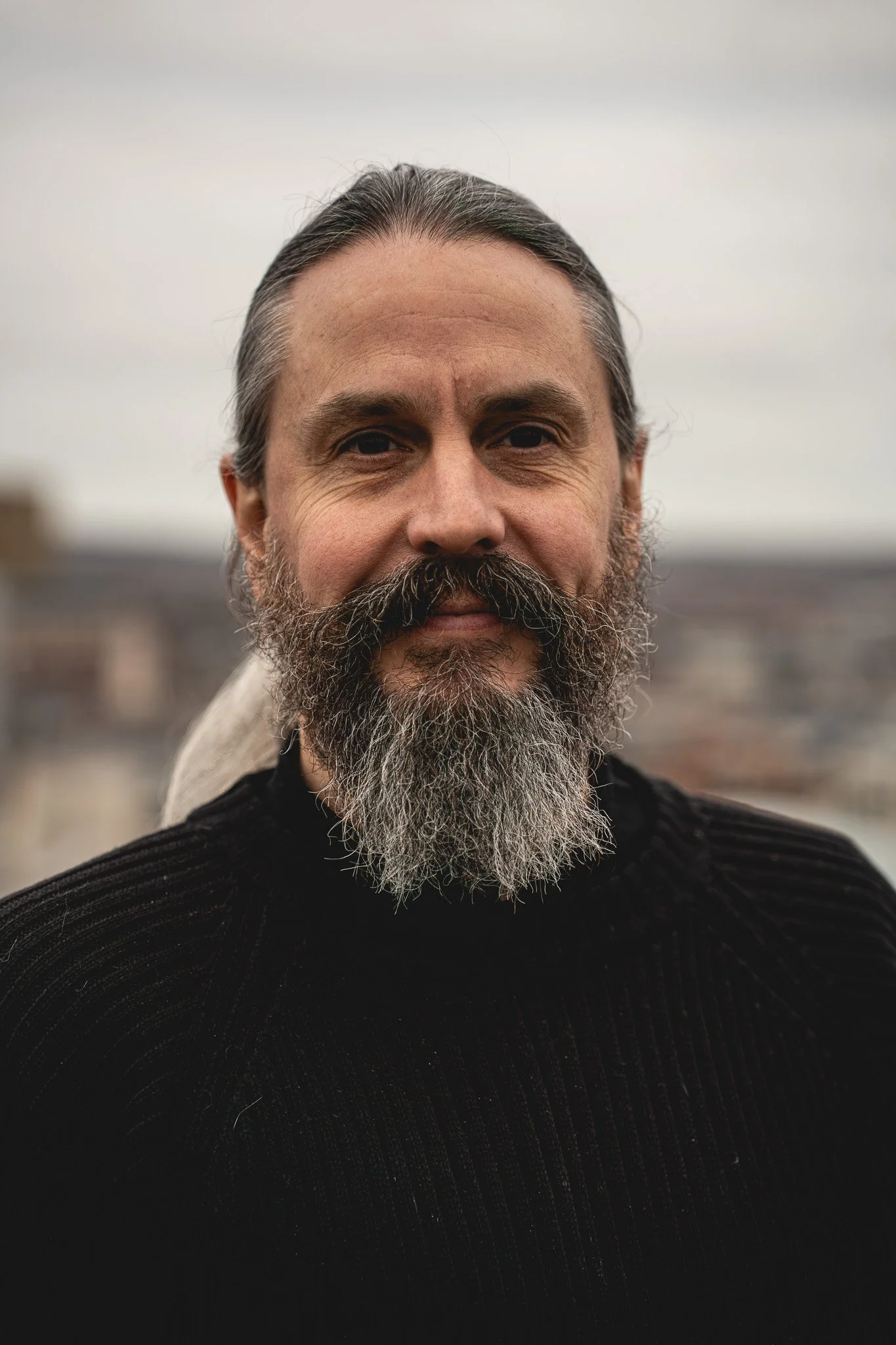 Tim Lewis, CTO and Co-Founder of Zygens, shown with trademark beard and long hair, wearing a black sweater, standing outdoors against a cloudy sky with a blurred Leeds cityscape in the background.