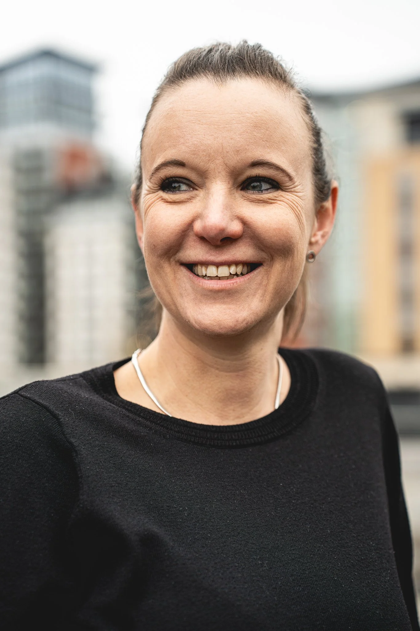 Close-up of a woman with a big smile, wearing a black top, small earrings, and a necklace, with a cityscape in the background.