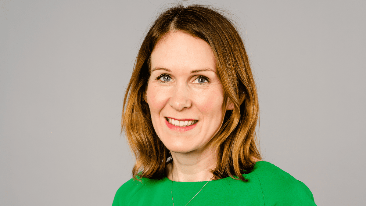 Ruth Handcock OBE wearing a green top and a necklace, smiling against a grey background.